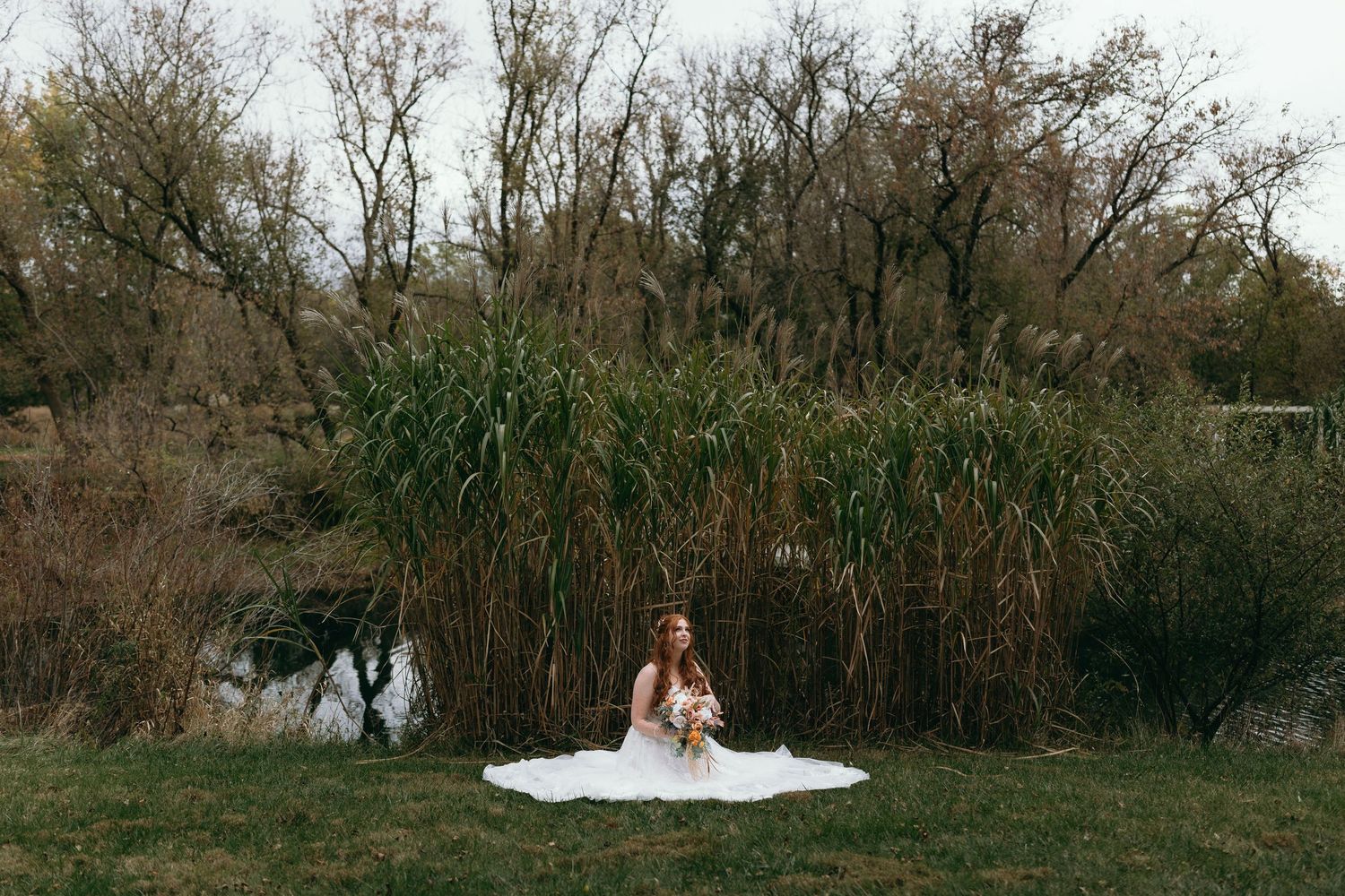 A figure in a white dress sits on the ground near a pond surrounded by tall reeds and bare trees in autumn.