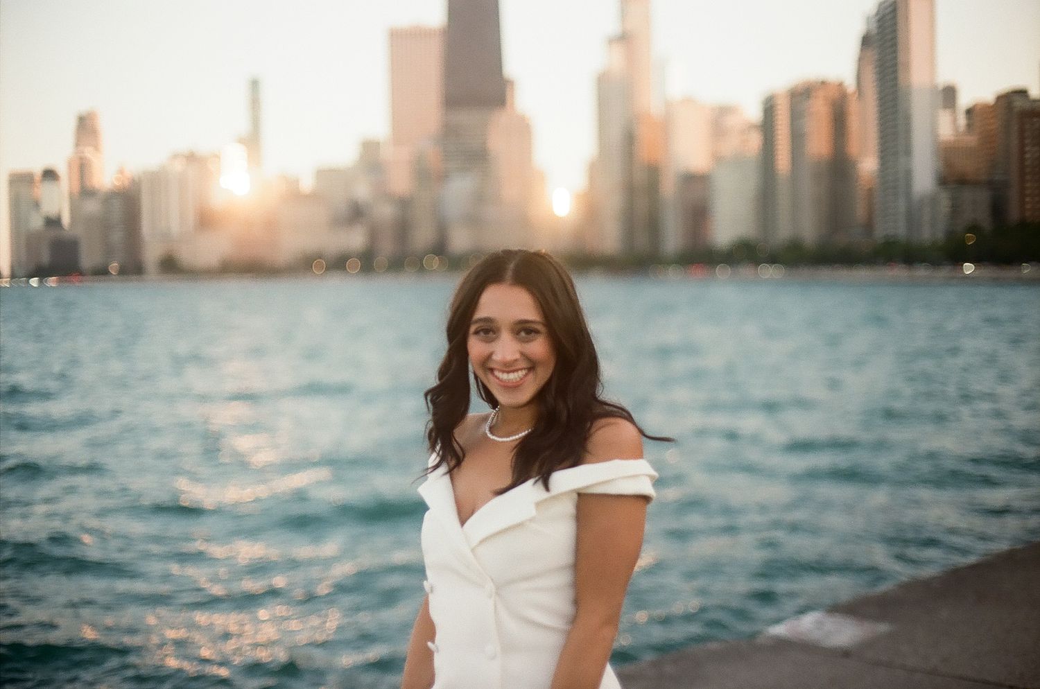 Woman in white dress smiles while sitting near urban waterfront at sunset with cityscape in background.