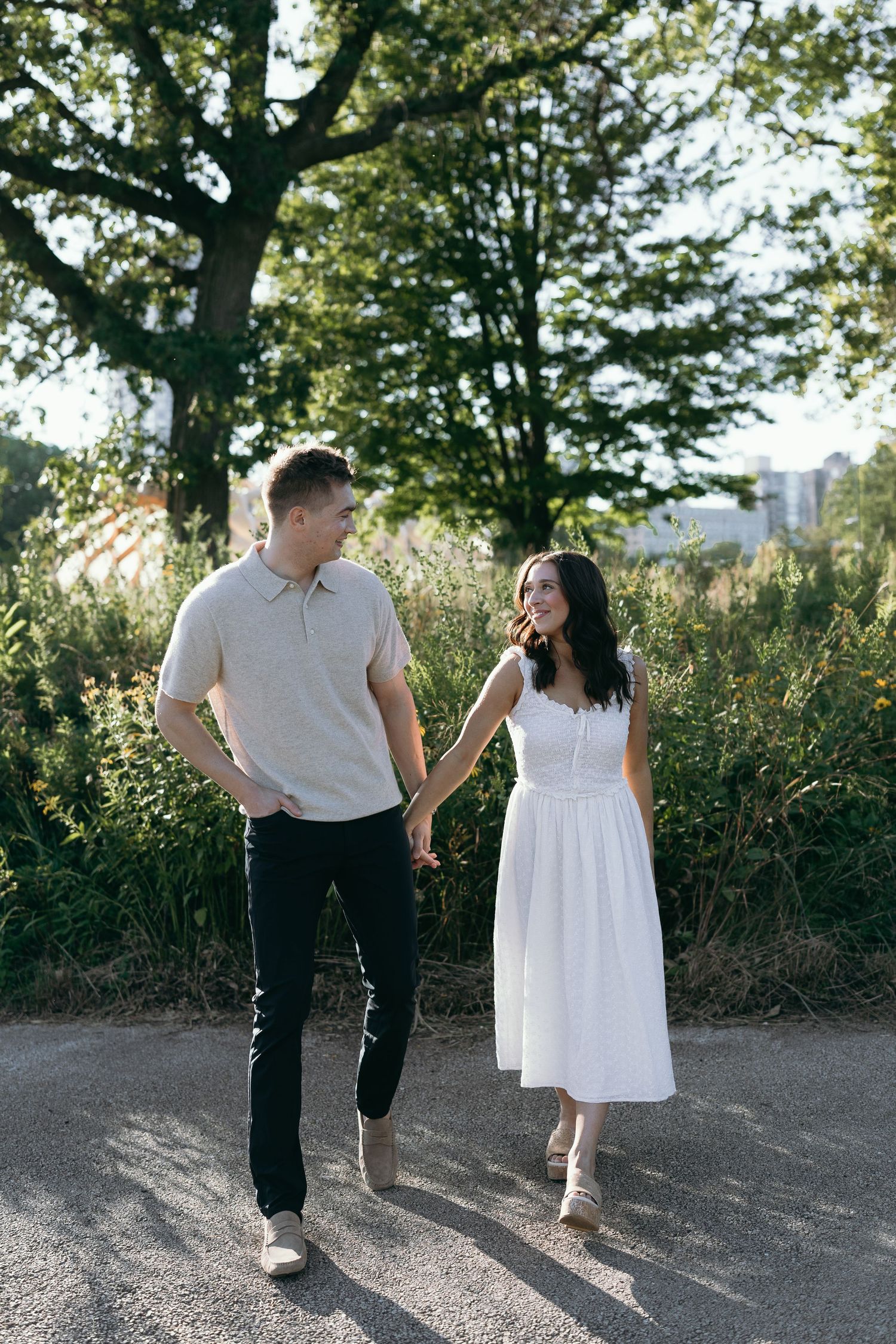 A couple walks together through a sunlit outdoor path surrounded by tall grass and trees on a summer afternoon.
