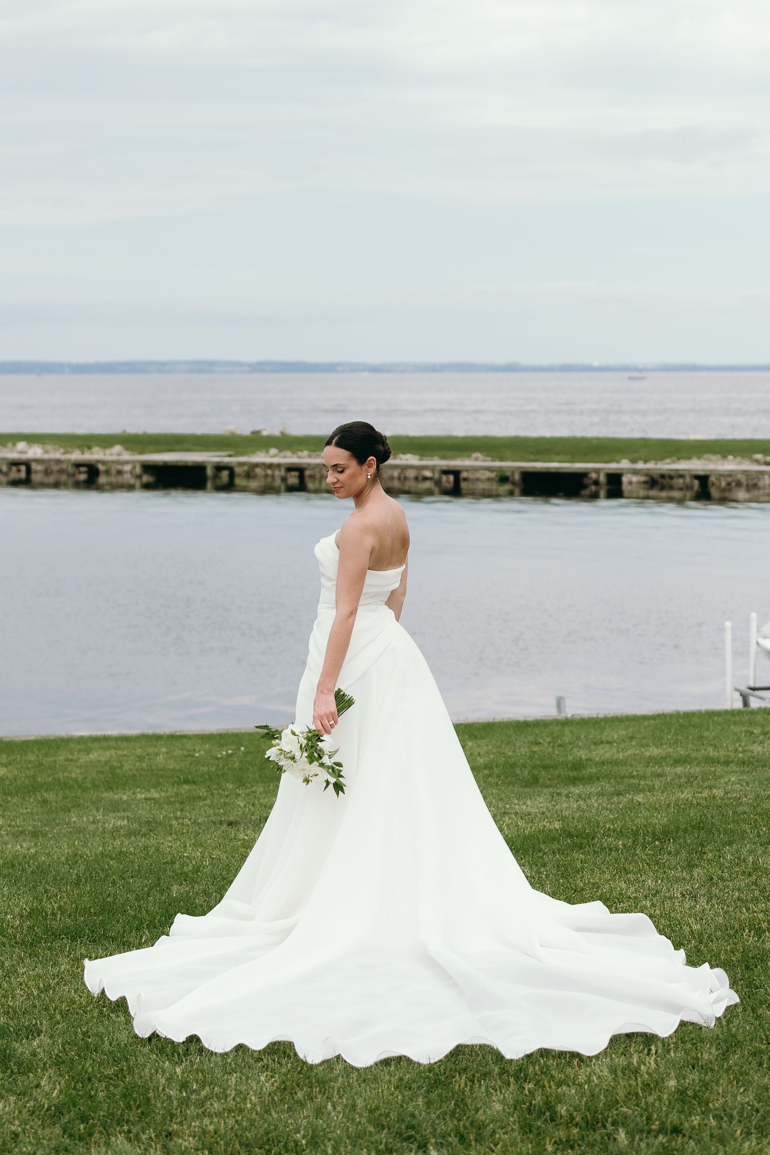 A wedding dress cascades elegantly across green grass with a serene waterfront backdrop.