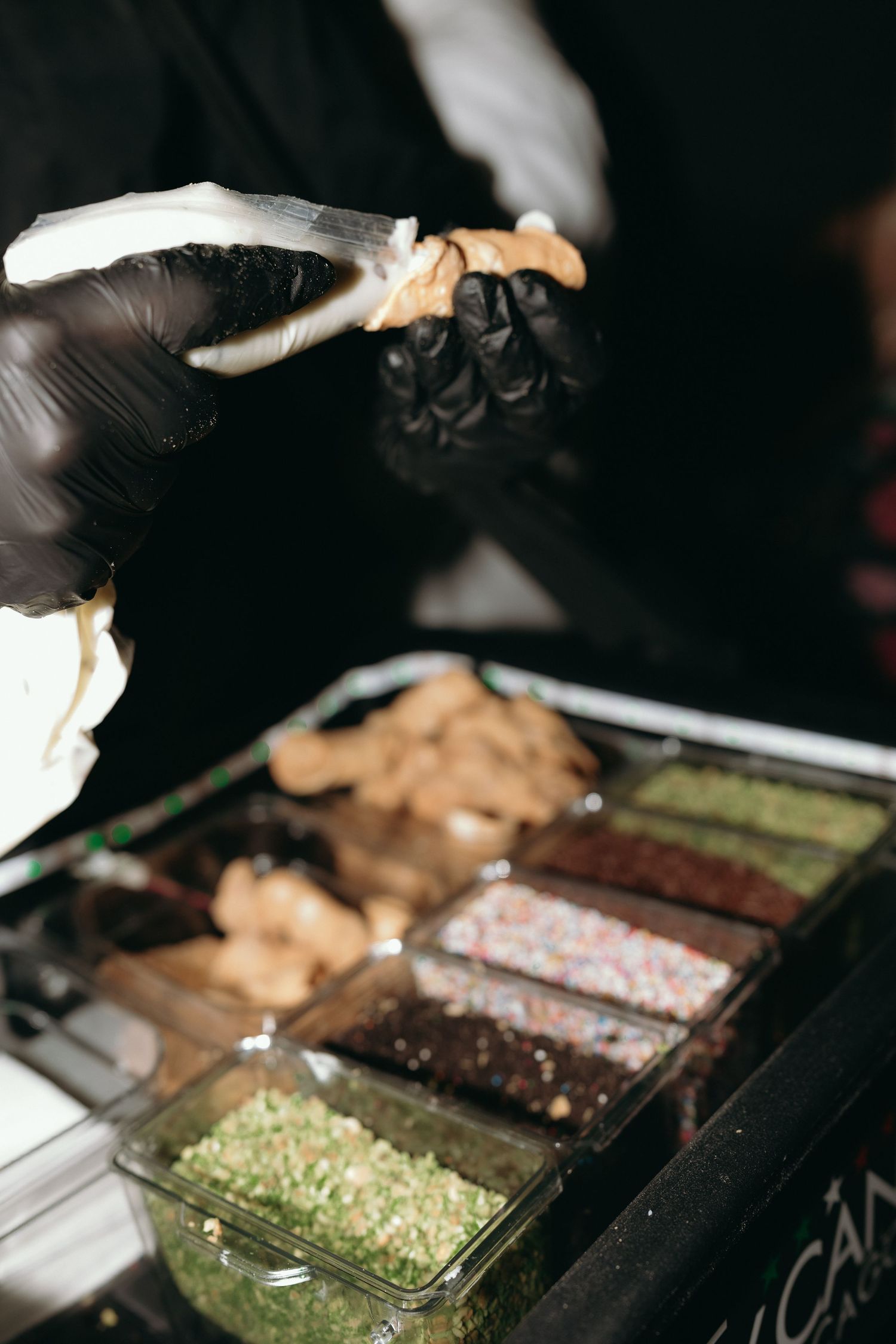 Caterer in black gloves preparing food at wedding reception dessert station with toppings.