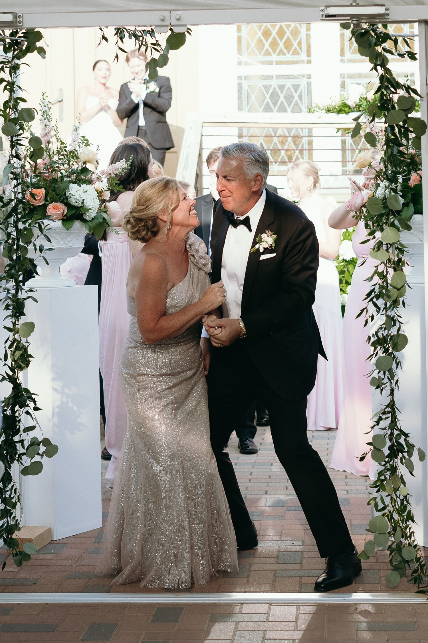 Elegant couple dancing under floral arch at wedding reception, woman in silver gown, man in black tuxedo.