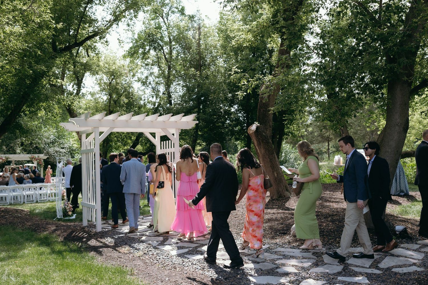 Outdoor wedding ceremony guests mingling near white arbor surrounded by lush green trees on a sunny day.