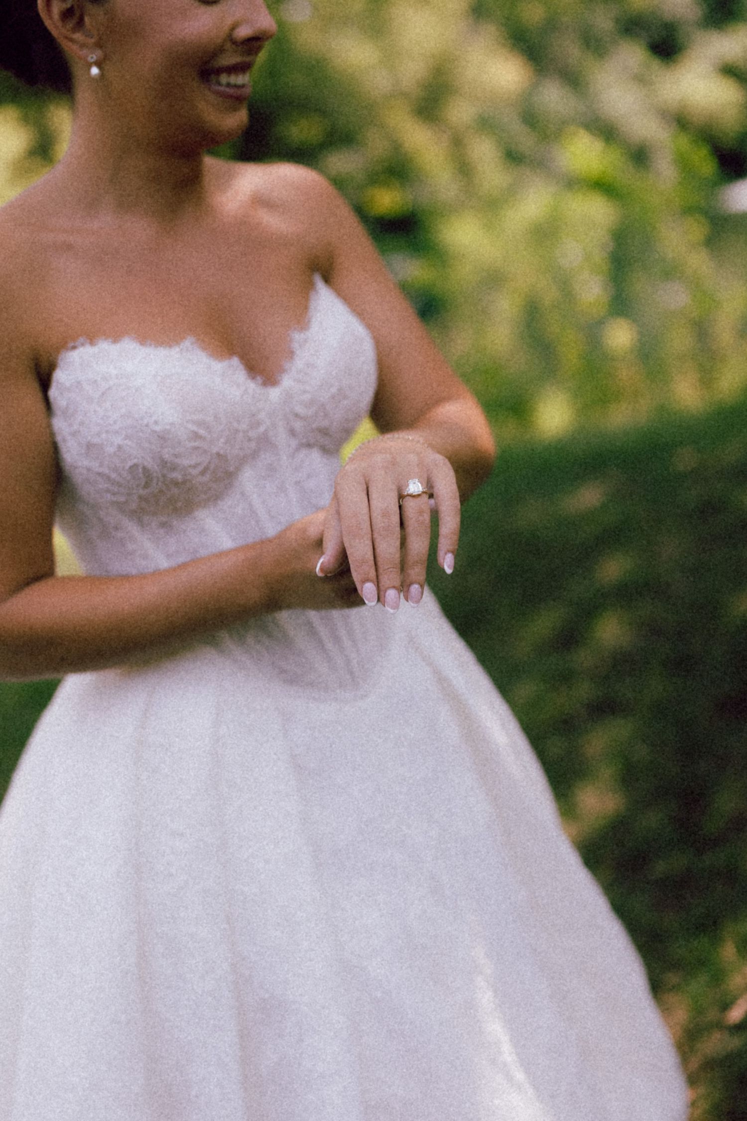 Smiling bride in strapless lace wedding dress displays elegant engagement ring in outdoor garden setting.