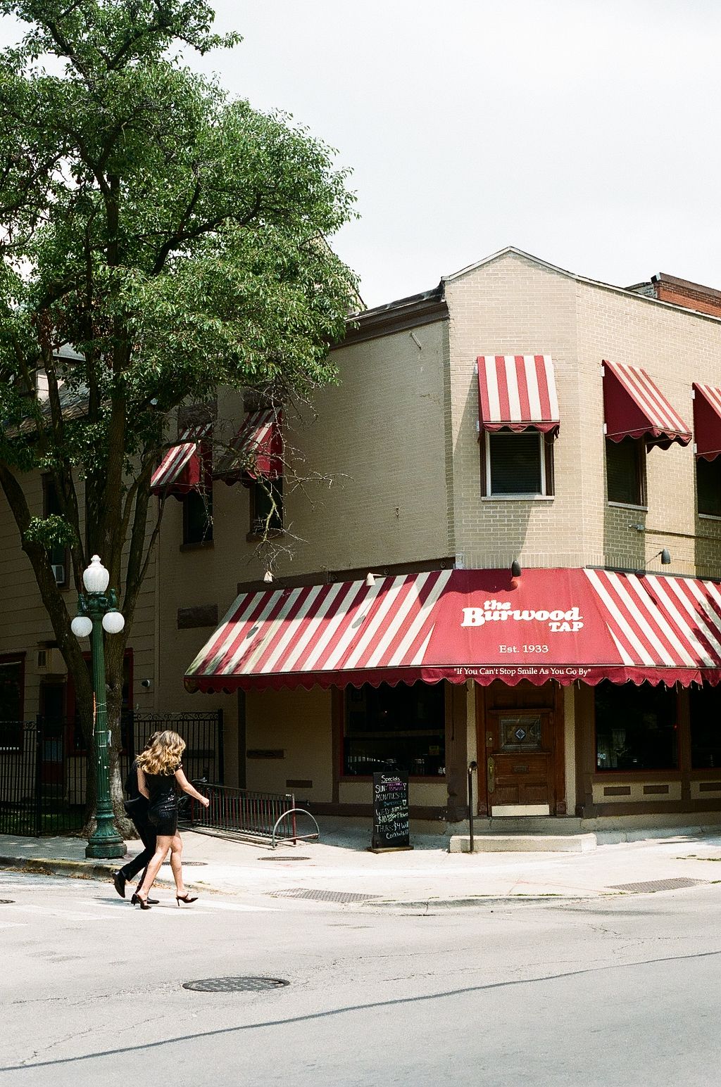 Woman walking past The Burwood Tap bar corner building with red and white striped awnings in urban neighborhood.