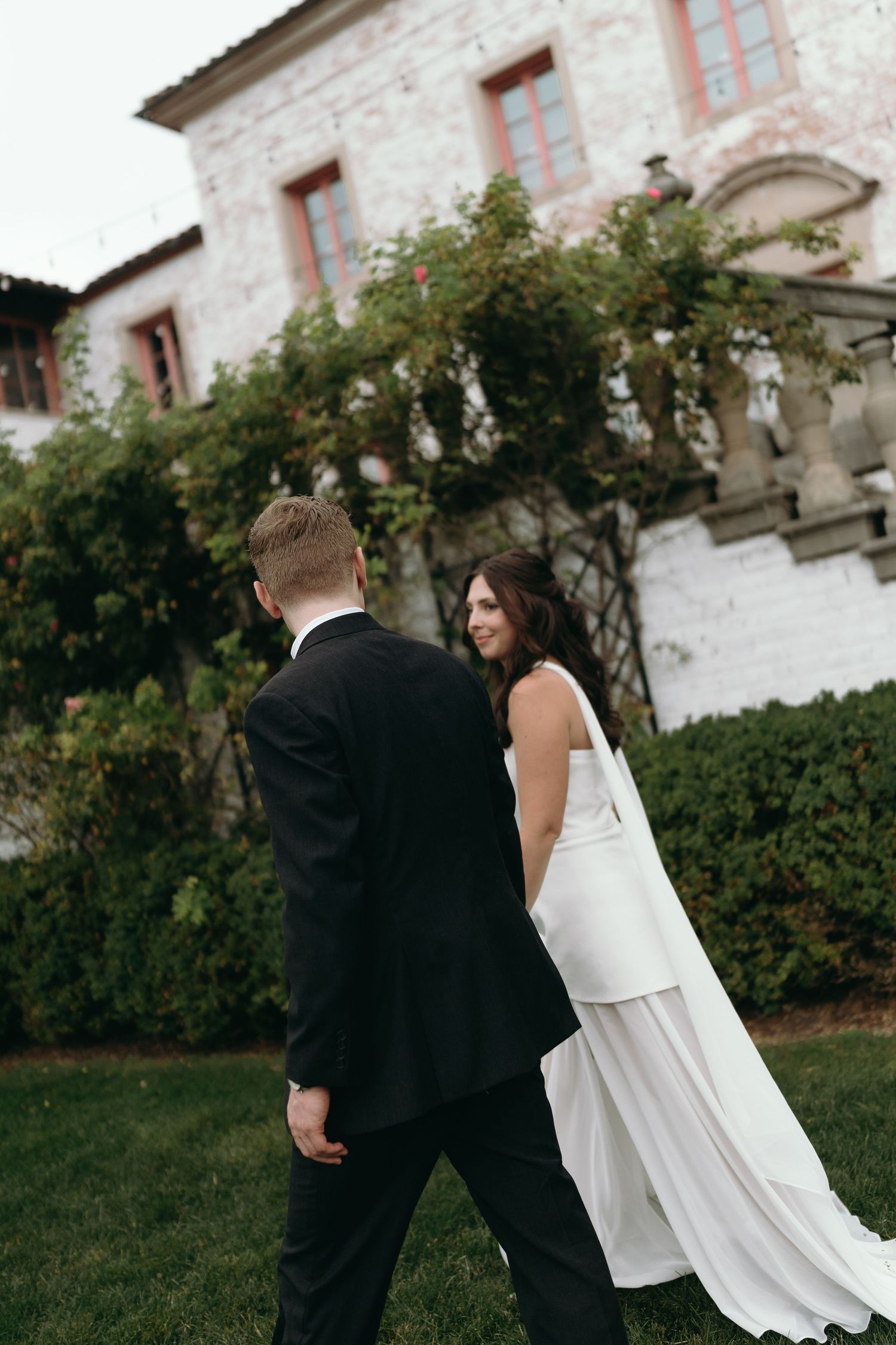 A bride in a flowing white dress and groom in black suit walk together on the lawn of a historic stone building.
