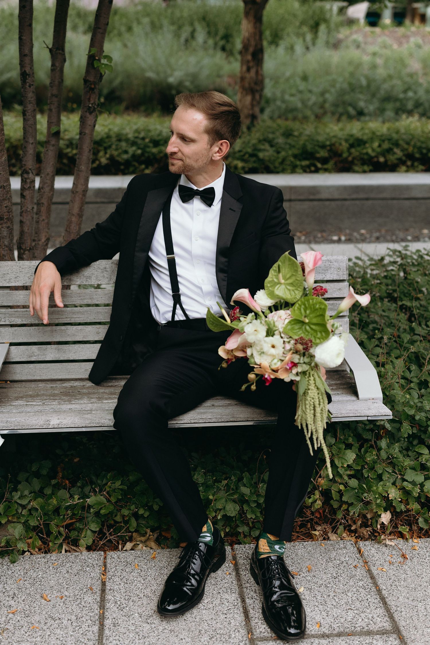 A person in a black suit sits on a bench holding a floral bouquet against a garden background.