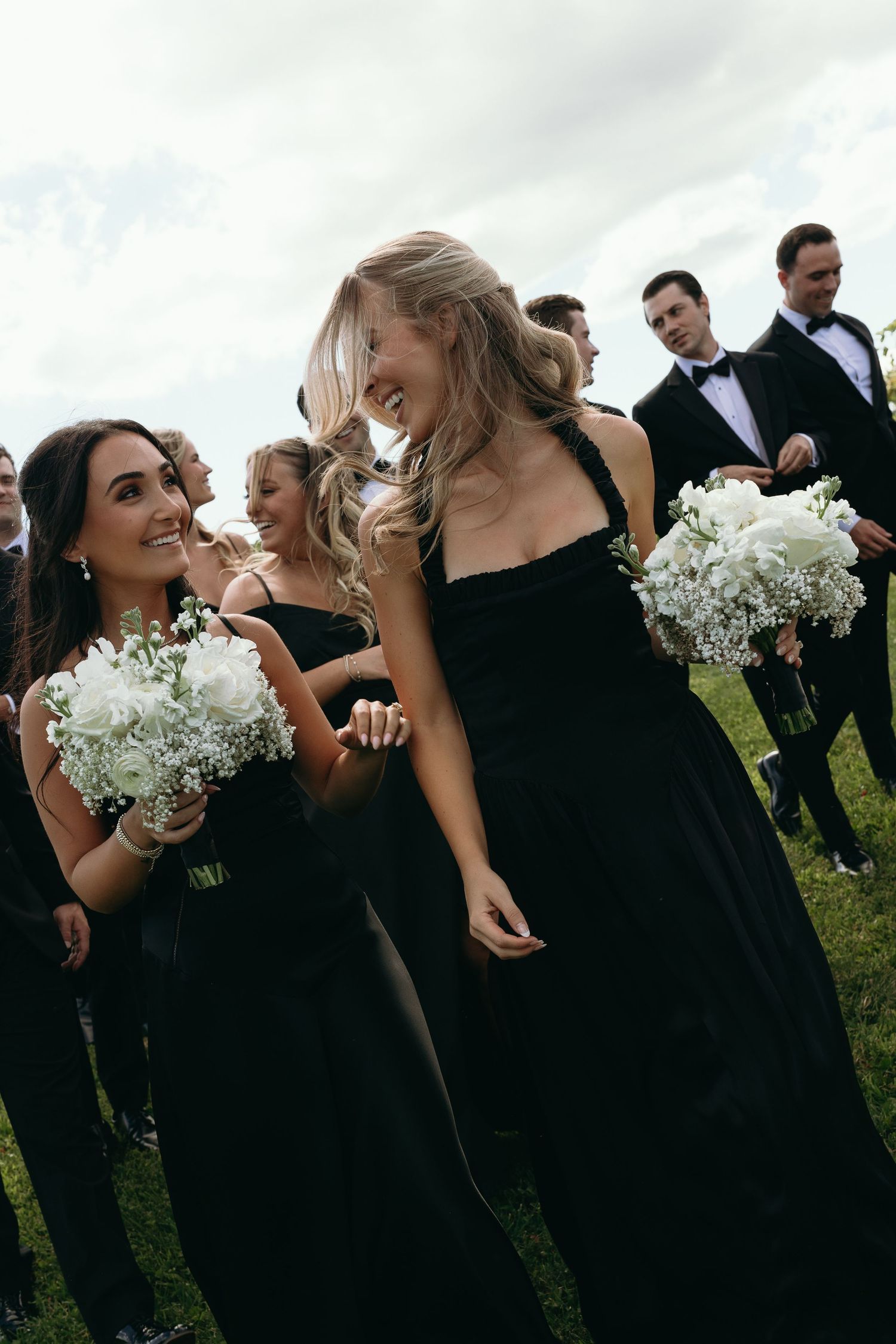 Wedding party members in black attire share joyful laughter while holding white bouquets outdoors.