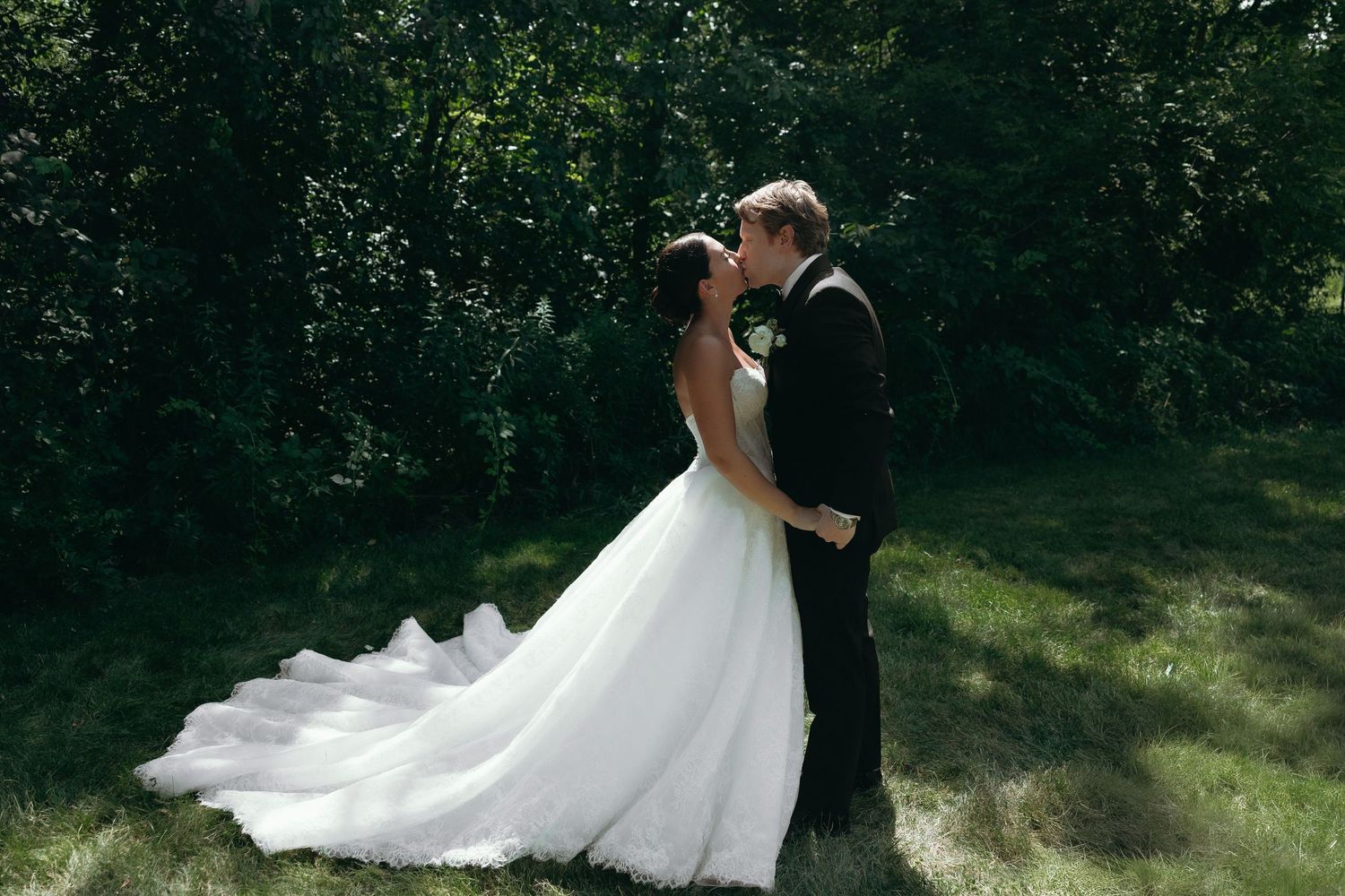 A bride and groom share a romantic kiss in a lush green garden, the bride wearing an elegant white ball gown with a long train.