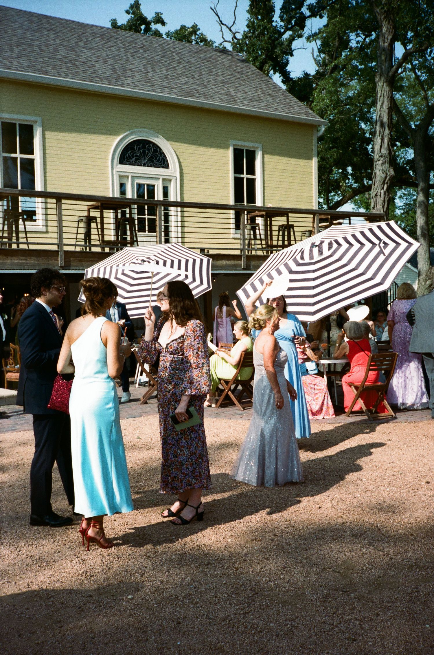 Wedding guests in elegant dresses gather outdoors under striped umbrellas near a yellow venue building on film