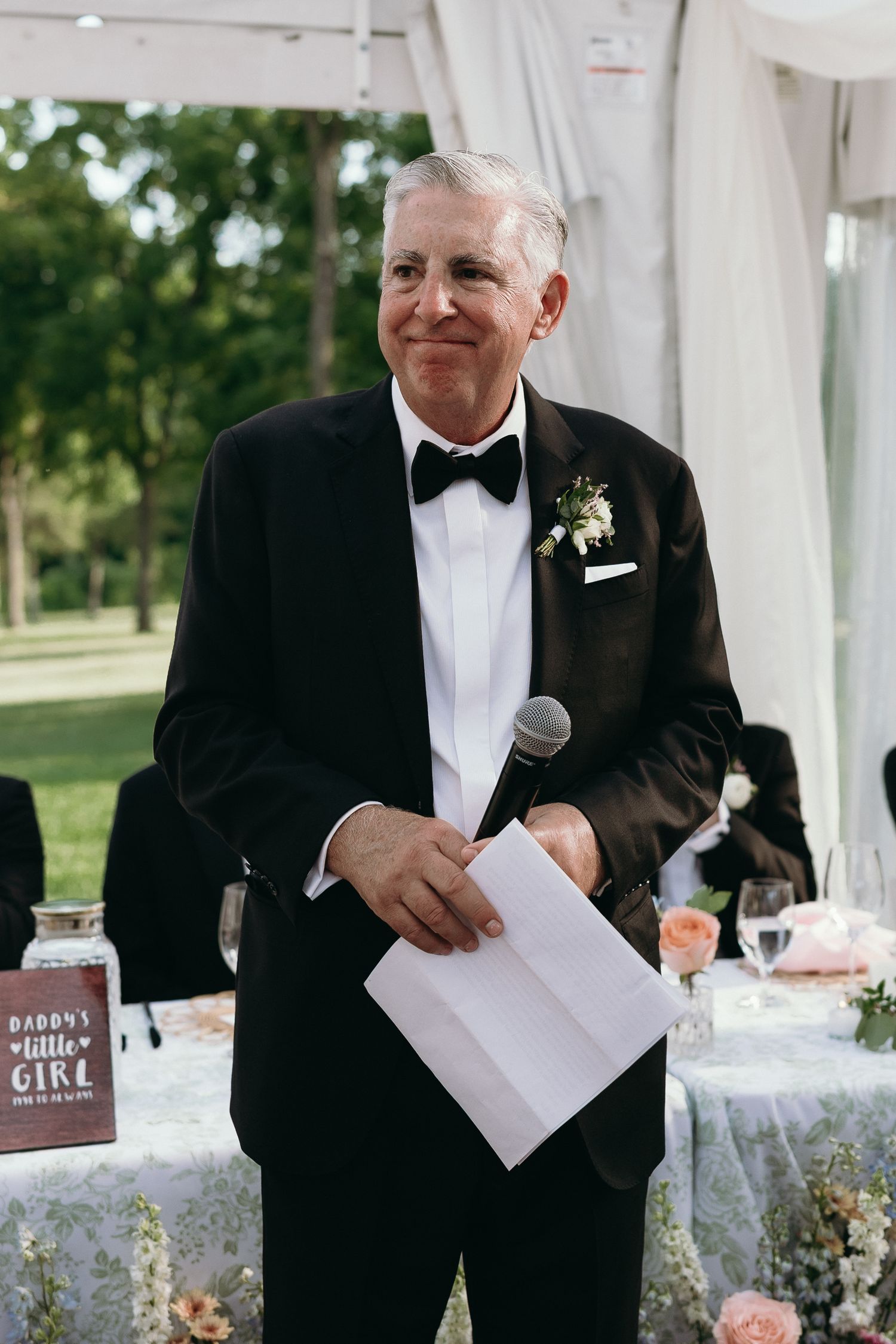 Father of the bride delivers a heartfelt speech holding a microphone and notes at wedding reception.