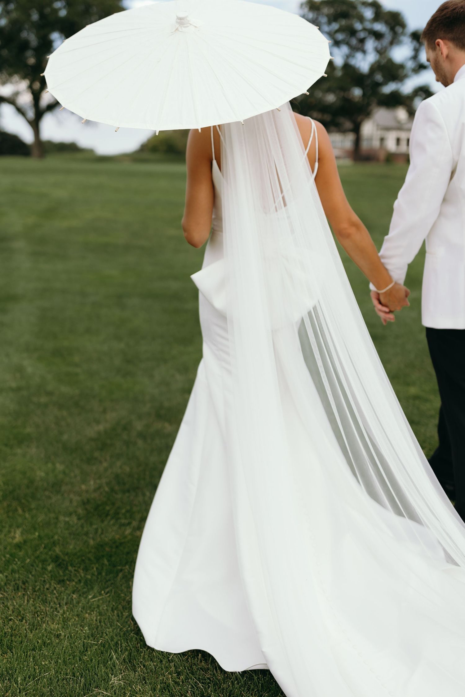 Elegant bride in flowing white gown holds parasol while walking with groom on manicured lawn.