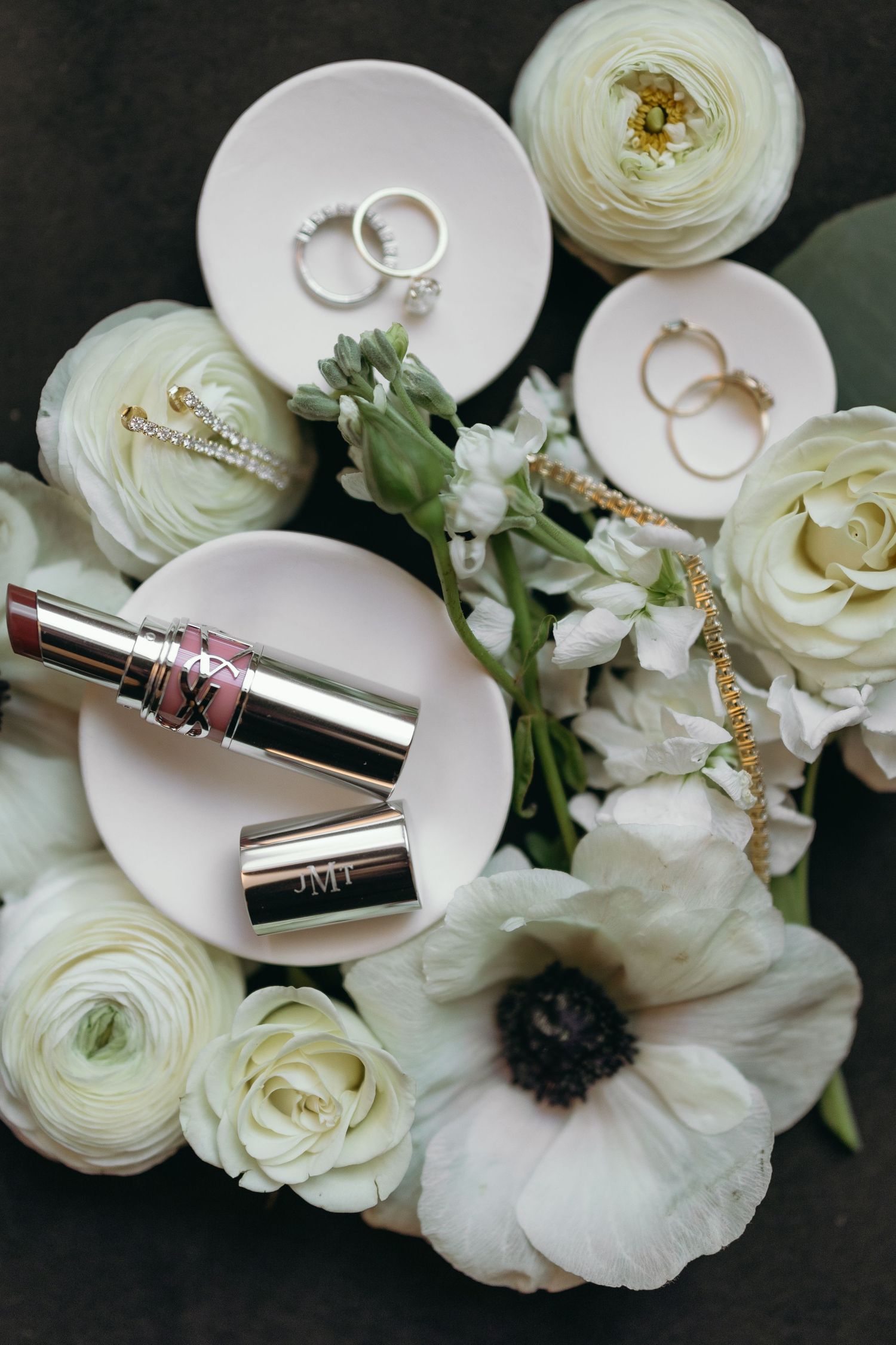 Wedding rings and lipstick arranged with white ranunculus flowers on dark surface.