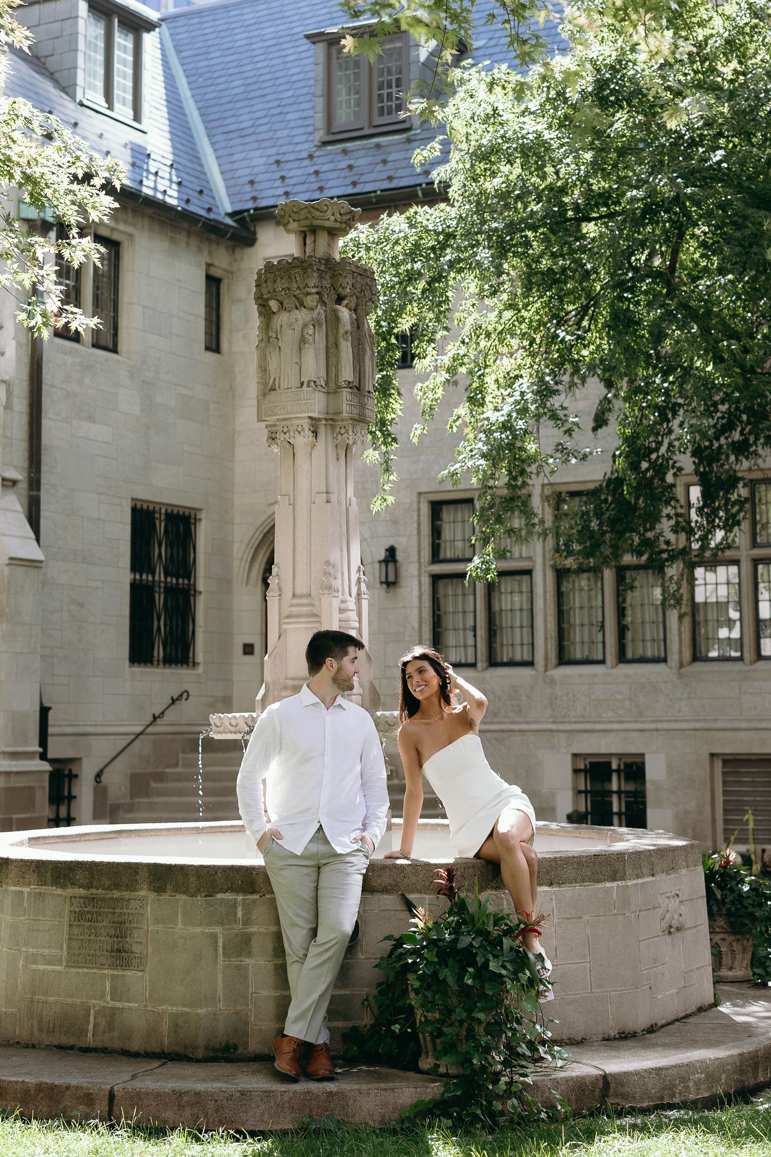 A couple sits together on a stone ledge in front of a Gothic-style building with climbing ivy and ornate architecture.