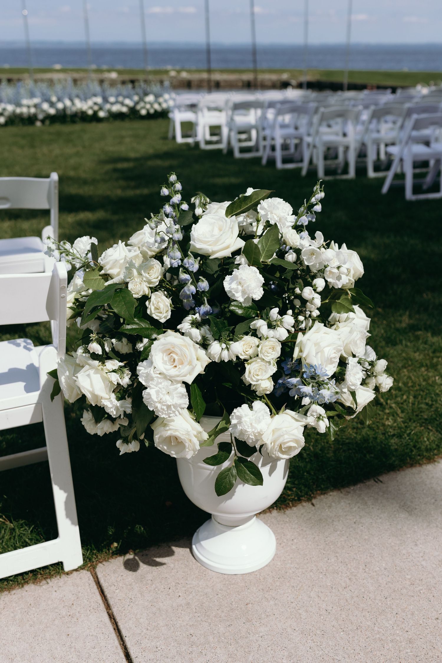 Elegant white rose and floral arrangement in white urn vase with ocean view and wedding chairs in background.