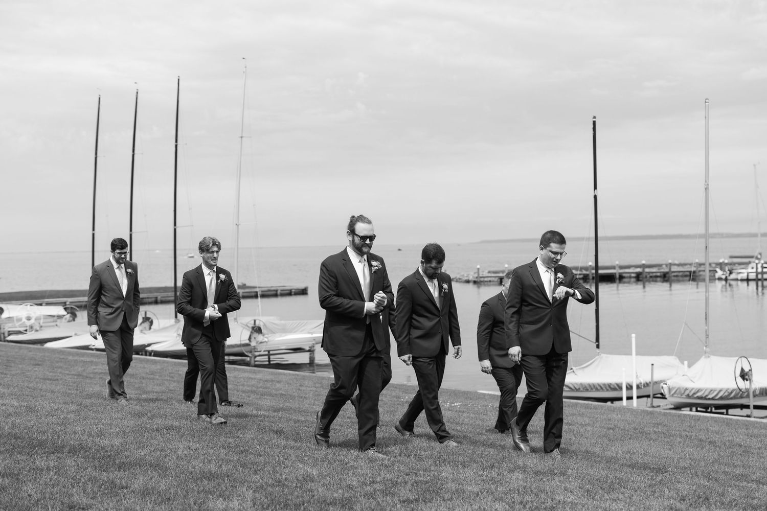 A black and white photo of groomsmen in suits walking together on a grassy area near a marina.