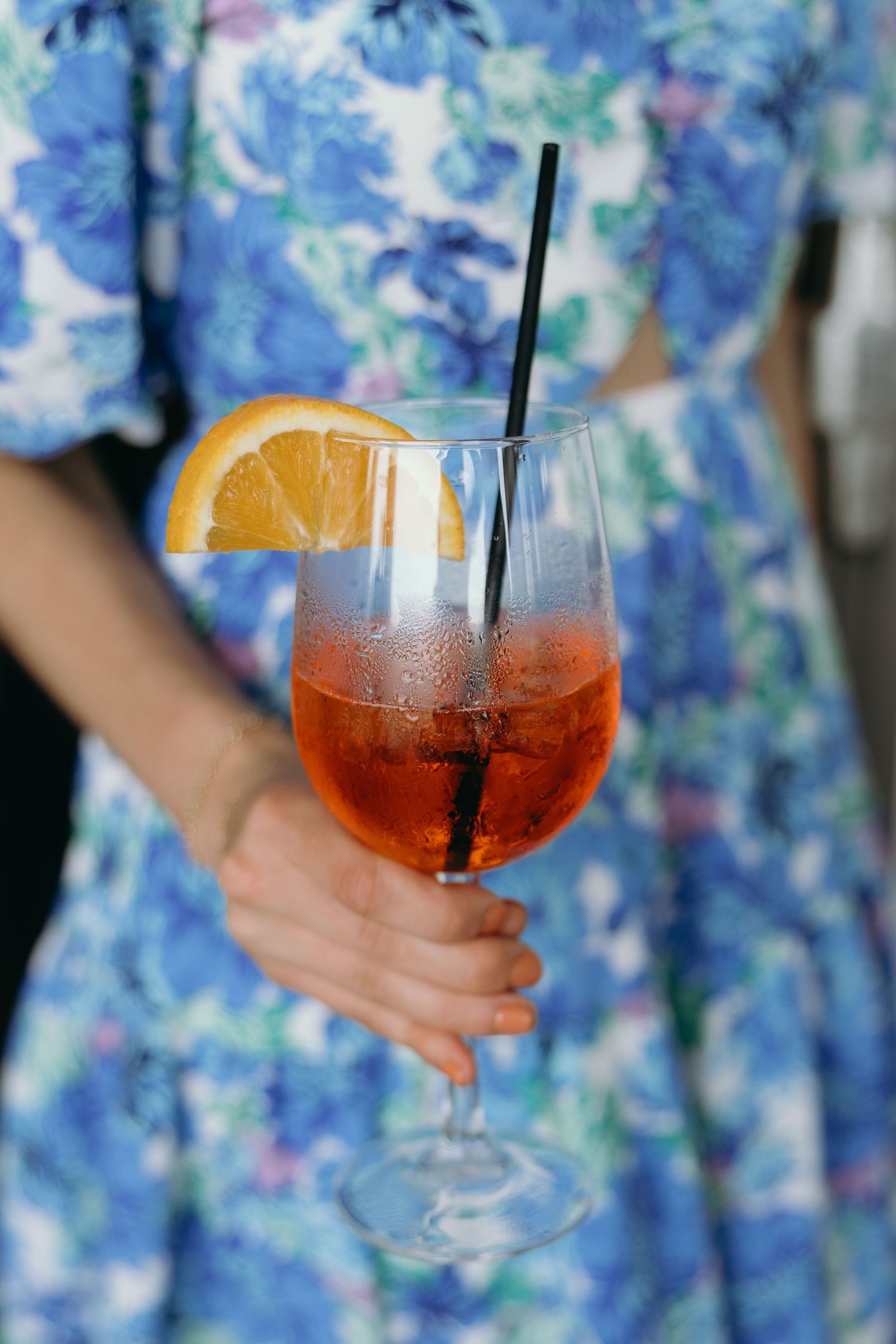 Hand holding a glass of Aperol Spritz cocktail garnished with orange slice against floral blue dress background.