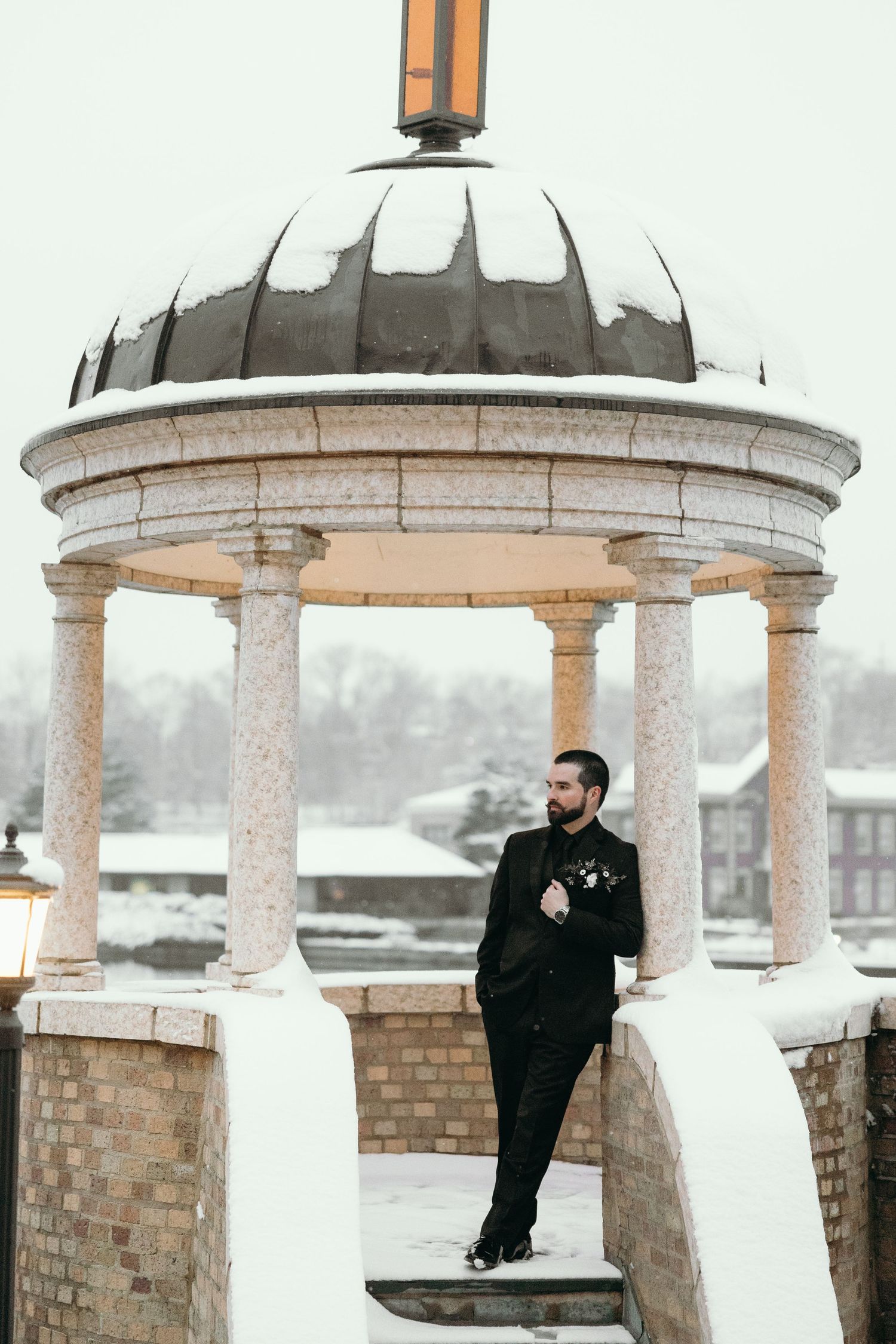 Groom in black suit poses in snow-covered classical gazebo setting.