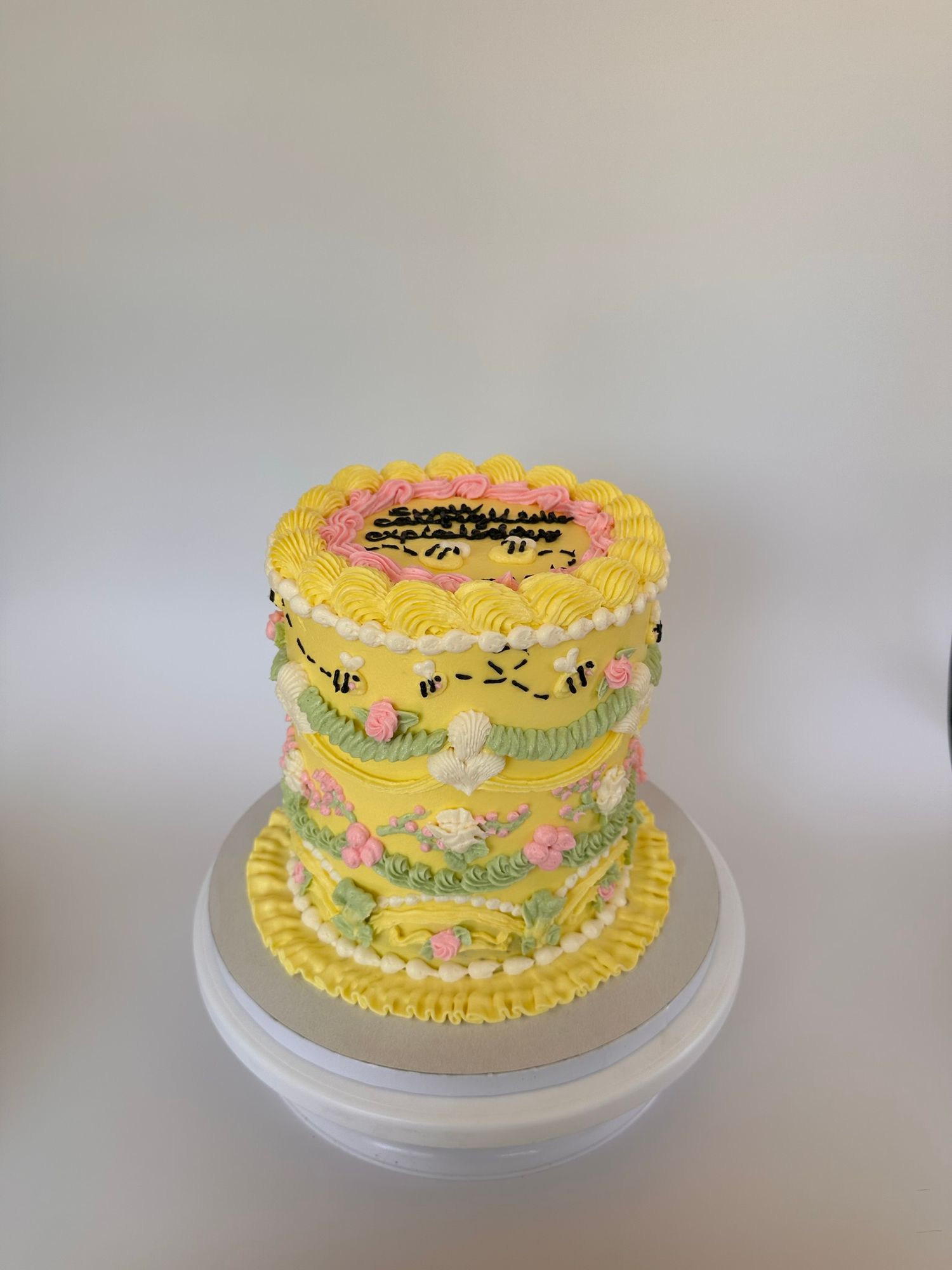 A yellow birthday cake decorated with pastel frosting flowers and piping details sits on a white cake stand.