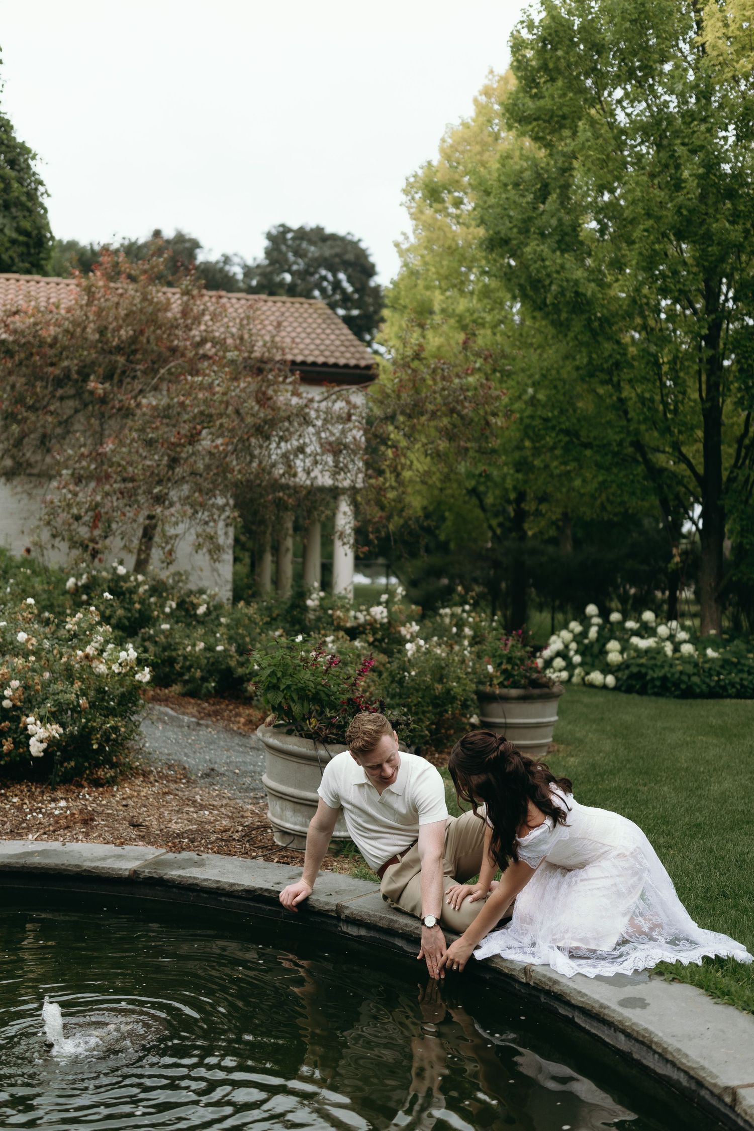 A couple in wedding attire sits by a garden pond near a white building with flowering bushes and tall trees in summer.