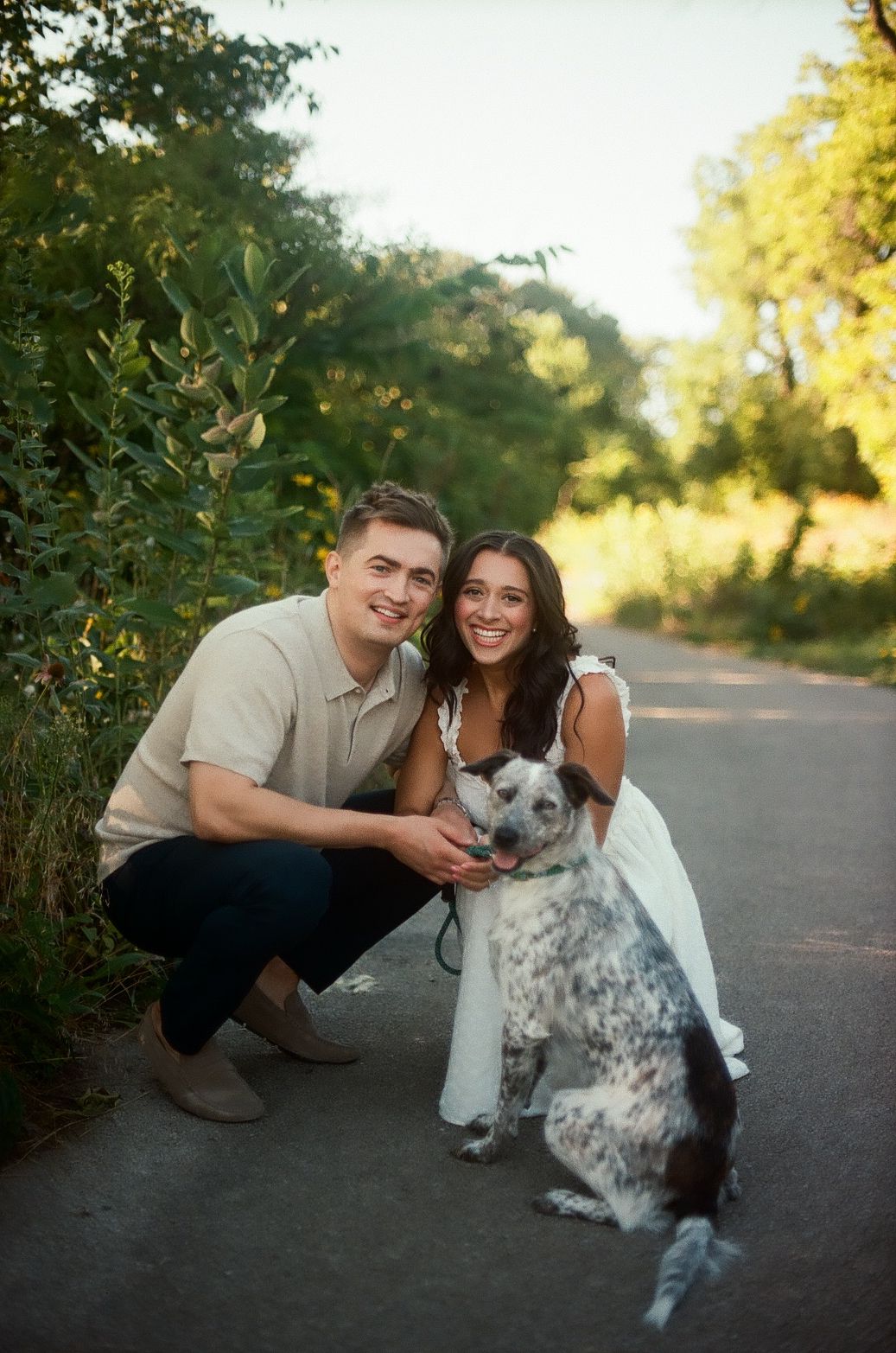 A couple and their spotted Australian Cattle Dog pose together on a sunlit path during a golden hour photo session.