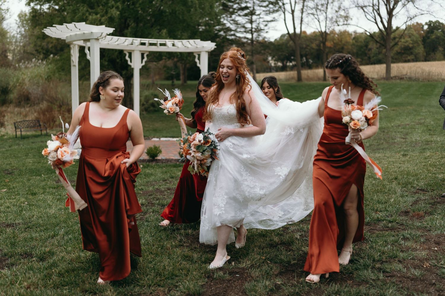 Wedding party in burgundy dresses and white gown pose playfully under garden arbor with floral bouquets.