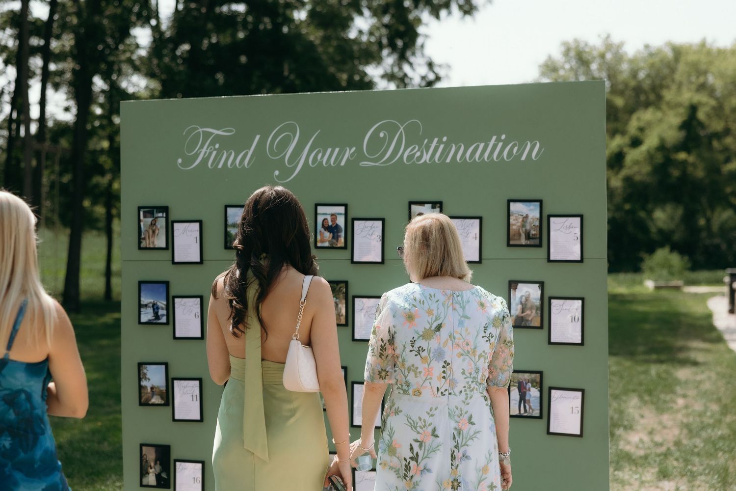 Wedding guests browse a sage green seating chart board titled Find Your Destination, displaying photos in an outdoor garden setting.
