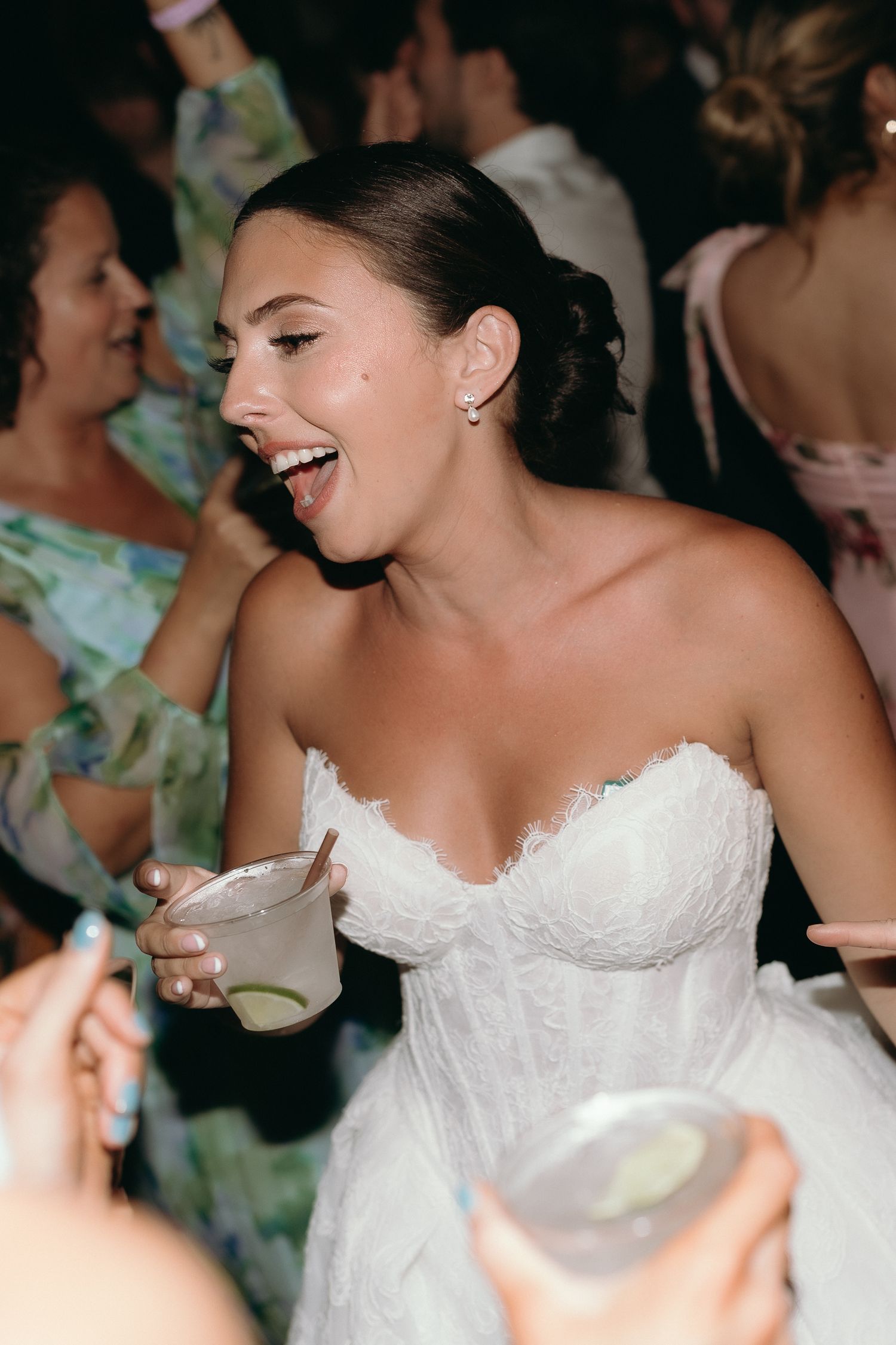 Joyful bride in a strapless white lace gown laughing and holding a cocktail at her wedding reception.