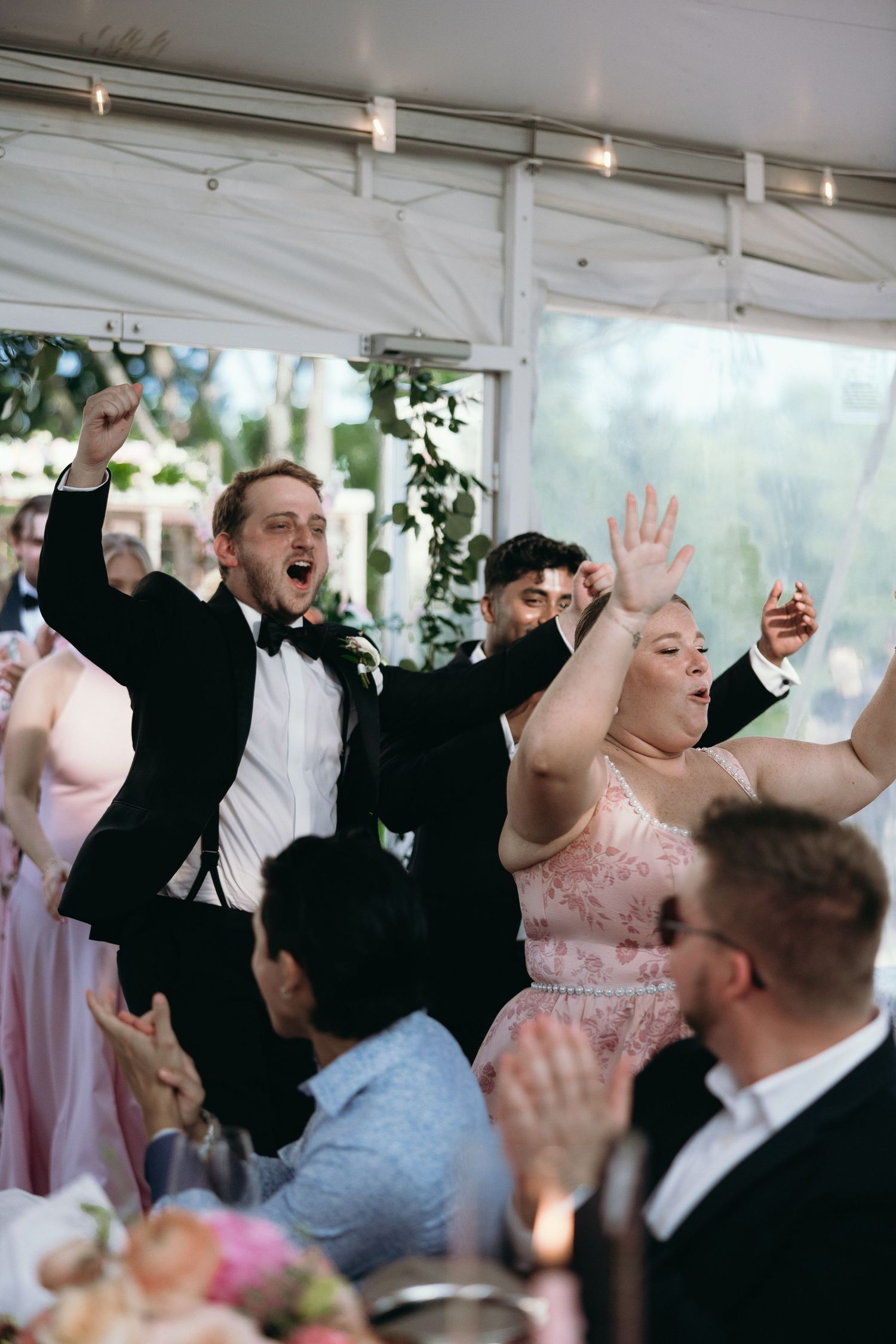 Excited wedding guests in formal attire cheering and raising hands during reception celebration under white tent.
