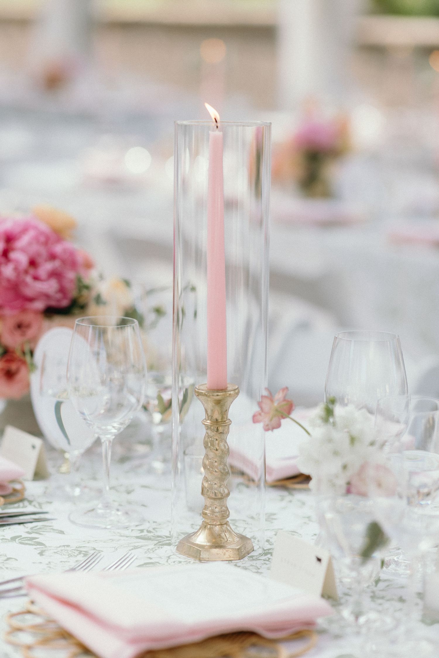Elegant wedding table setting with lit pink taper candle in gold holder, crystal glasses, and pink floral arrangements.