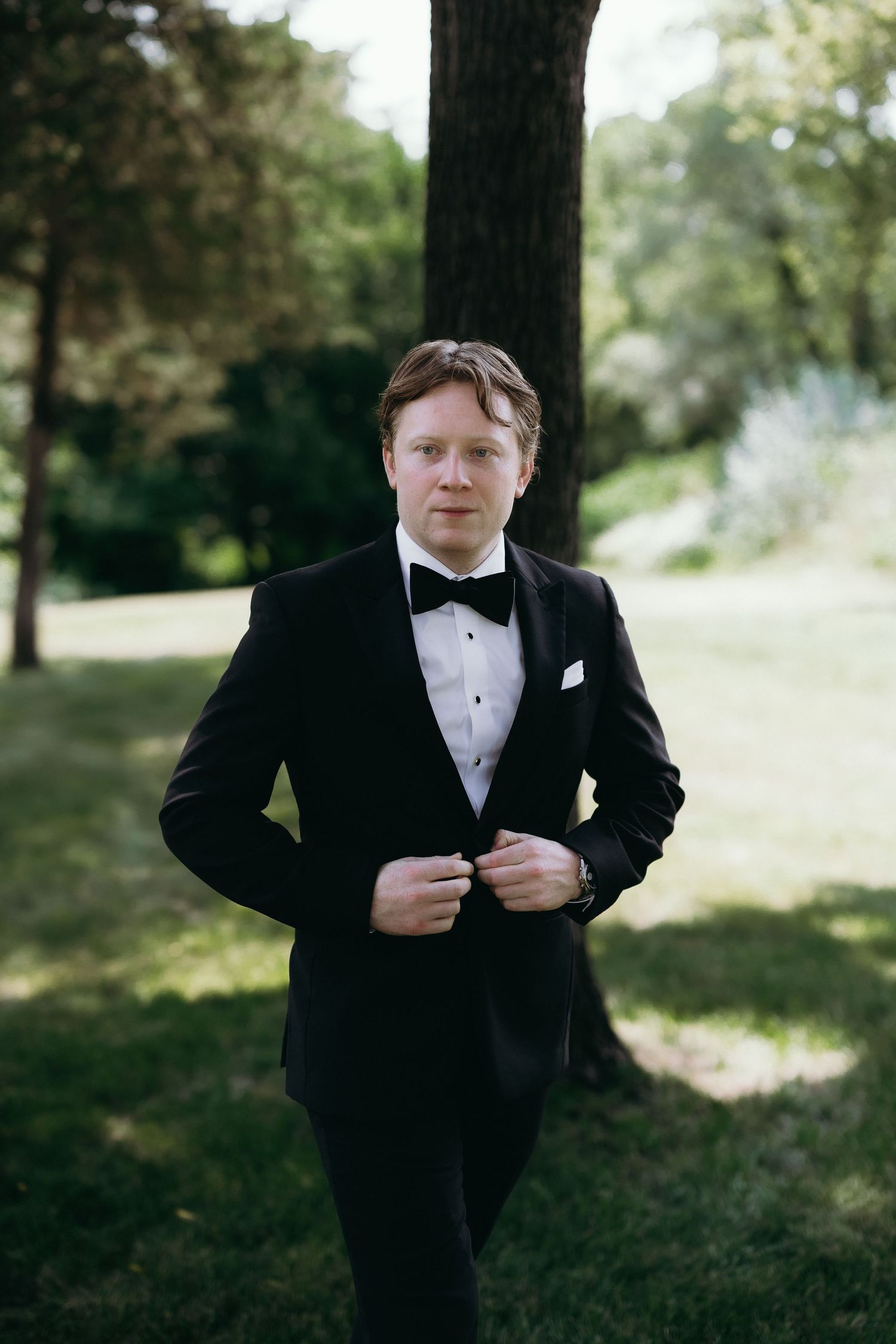 Groom in black tuxedo with bow tie adjusting jacket buttons outdoors in lush green park setting.