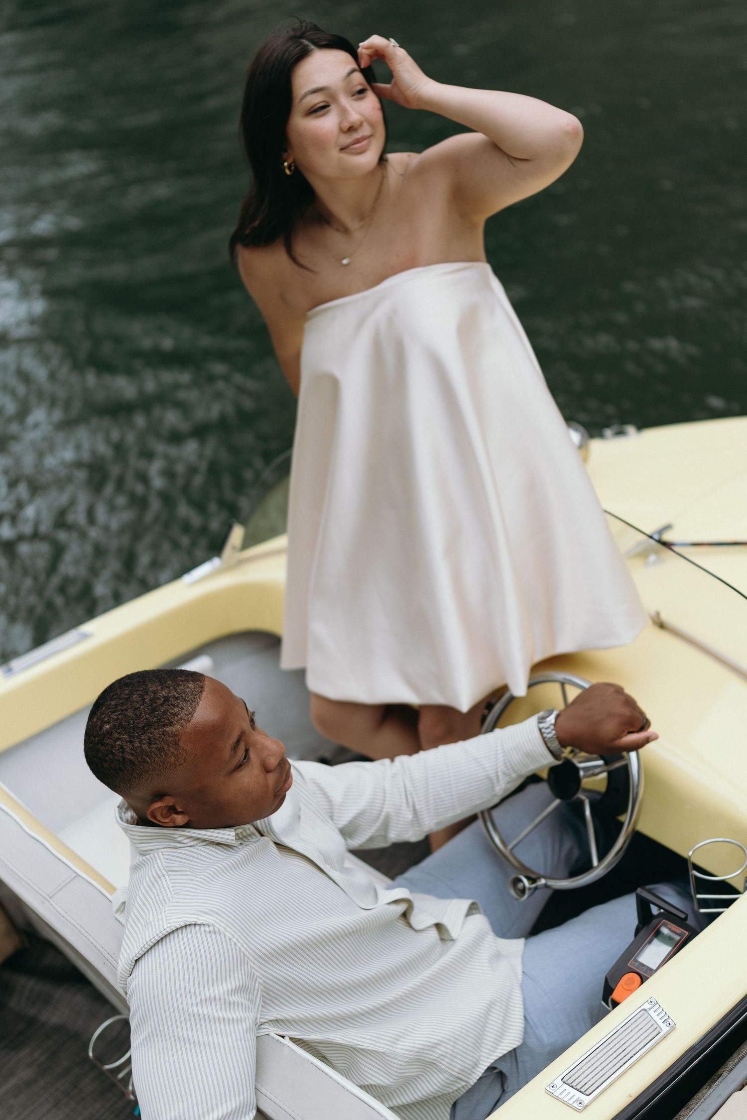 A couple on a vintage boat, woman in white strapless dress standing while man steers on calm water.