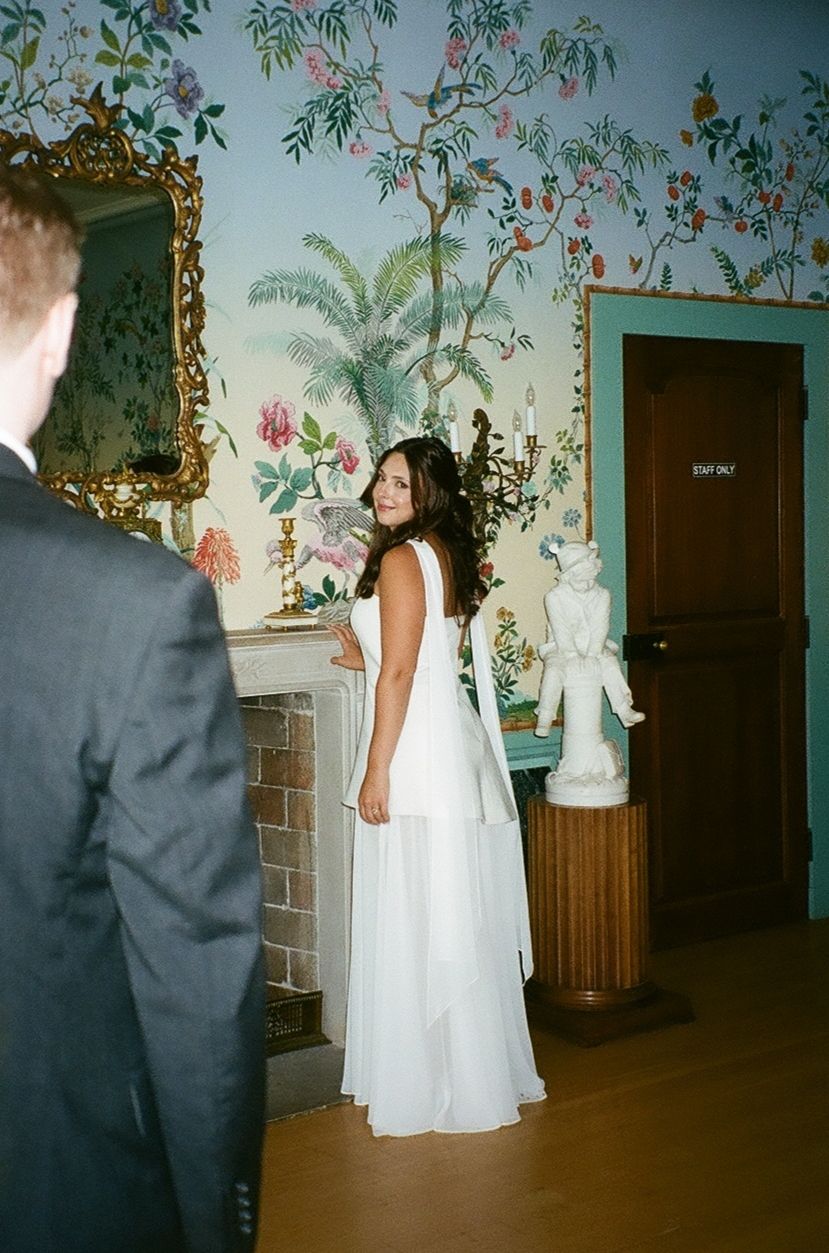 Someone in a white wedding gown poses in front of ornate floral wallpaper with a gold-framed mirror and decorative statue.