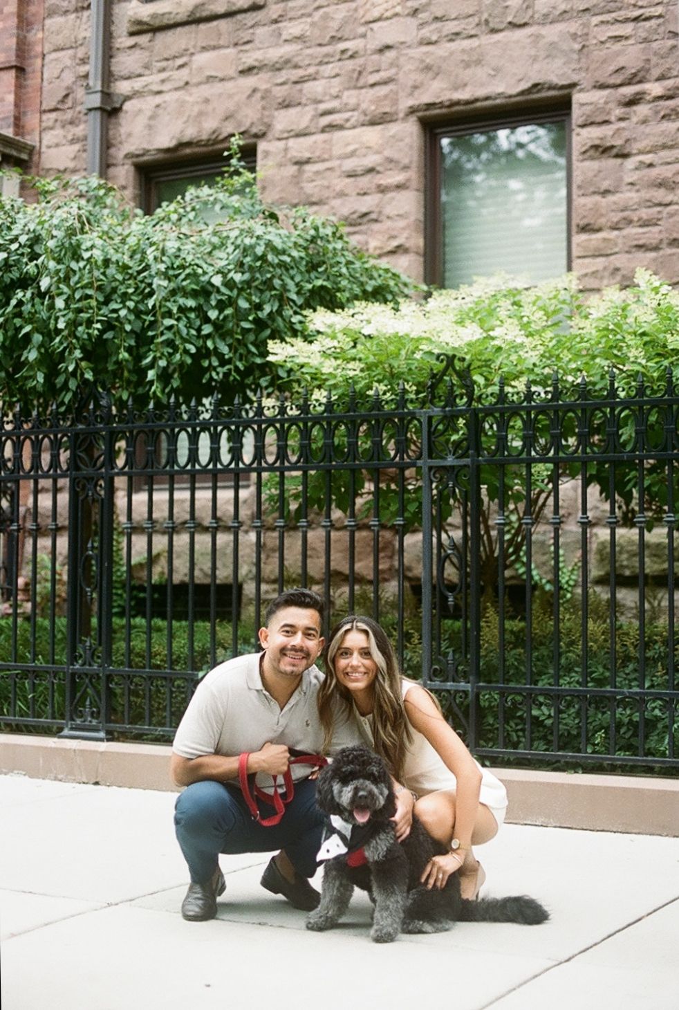 Three progressive photographs of a couple posing with their black dog on a sidewalk near a brick building with an iron fence.