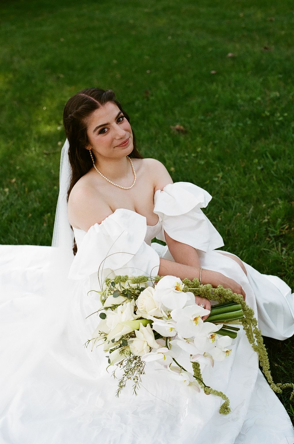Bride in white strapless gown sitting on green grass holding elegant white orchid bouquet with pearl necklace.