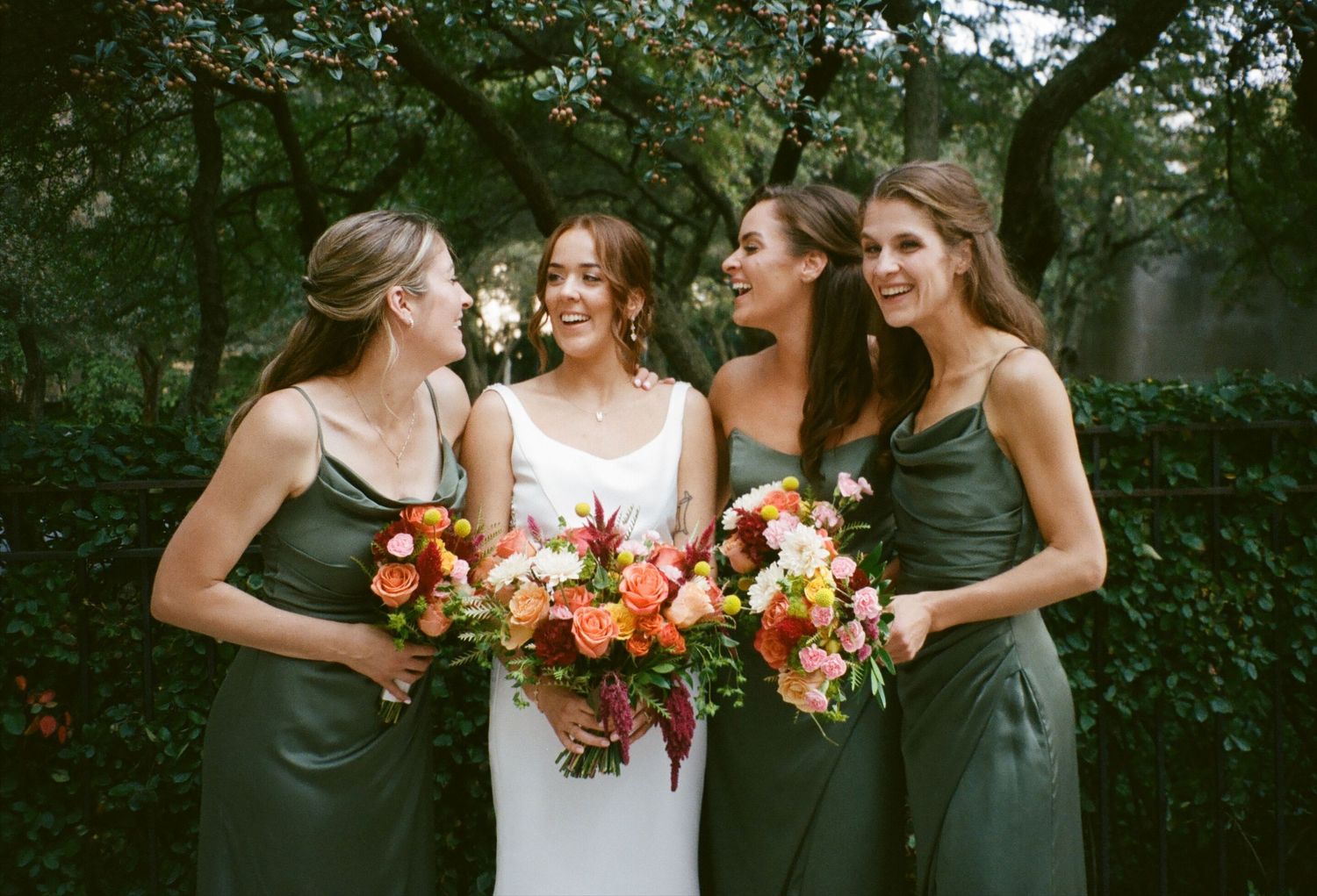 Bride in white gown with three bridesmaids in dark green dresses holding vibrant orange and pink bouquets outdoors.