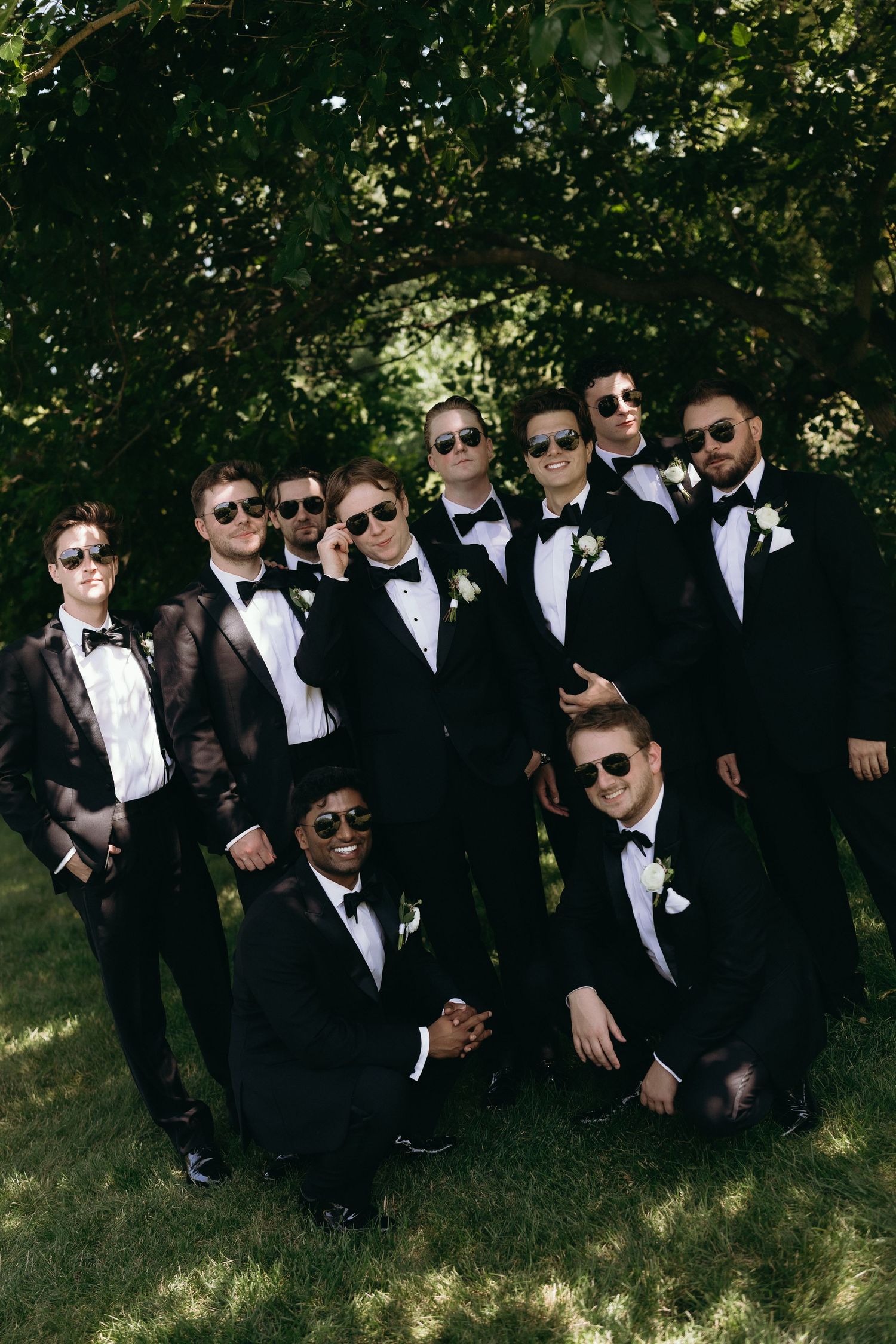 Groomsmen in black tuxedos and sunglasses pose together outdoors under green trees at a wedding.