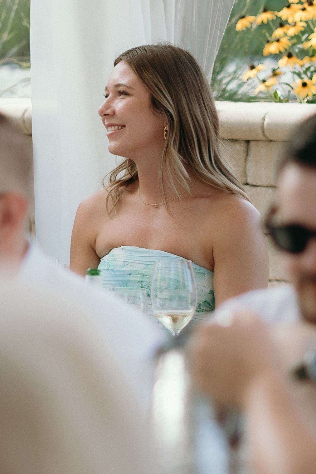 Smiling woman in strapless green dress holding wine glass at outdoor event with yellow flowers.