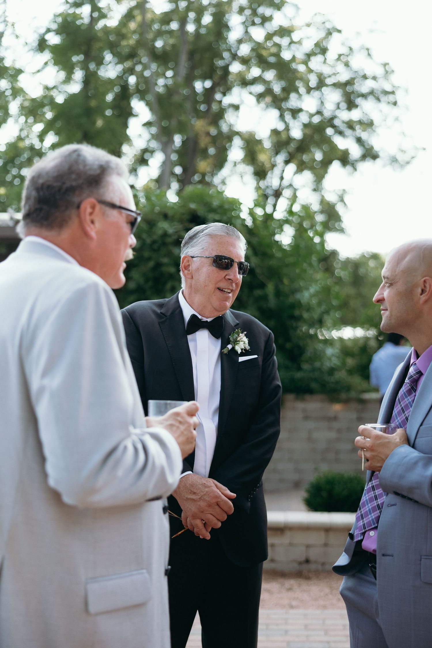 Three men in formal suits share a laugh outdoors at a wedding reception on a sunny day.
