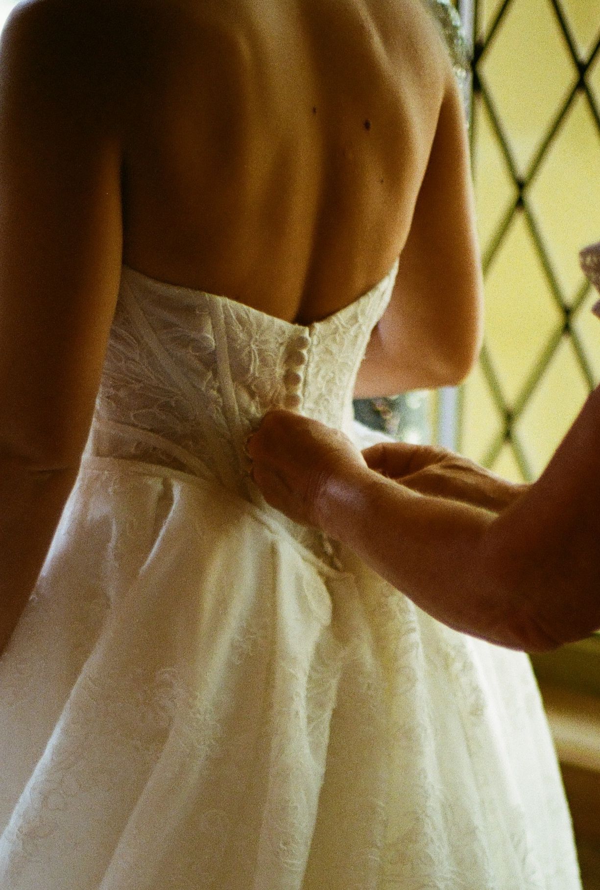 Bride in white lace wedding dress being buttoned up from behind near a stained glass window.