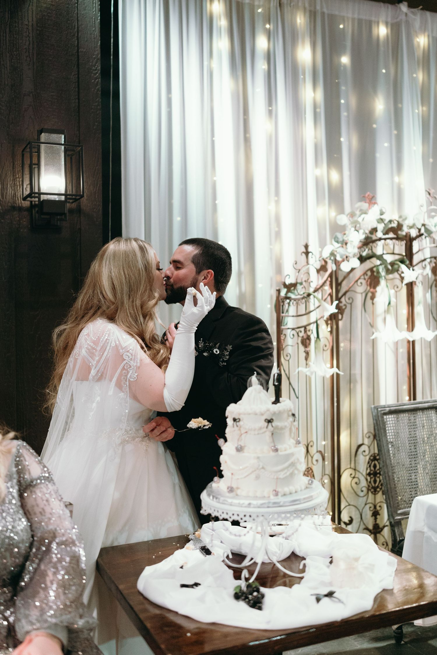 Elegant white wedding cake display with twinkling fairy light backdrop during a romantic reception moment.