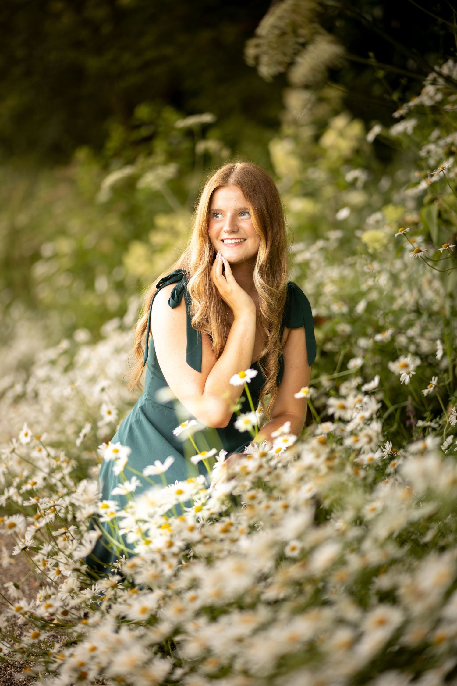 Young woman in green dress sitting among white daisies in a sunlit garden, smiling warmly.