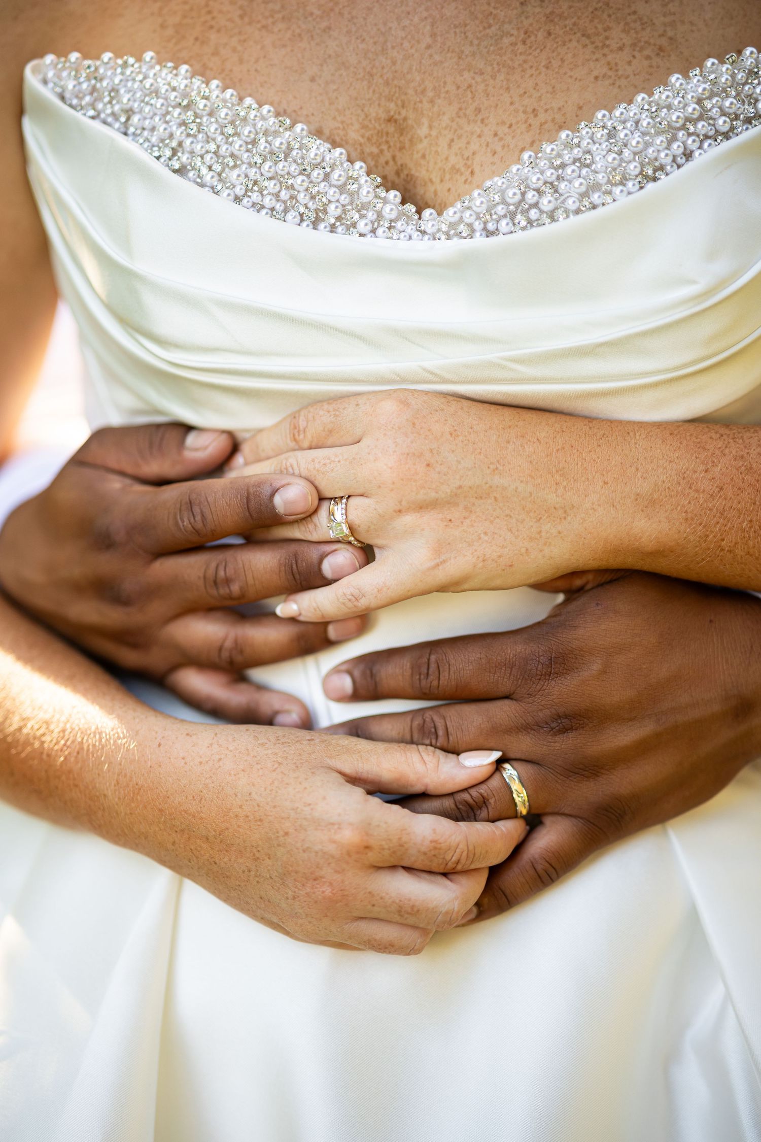 Newlywed couple embracing, showing gold wedding rings on their hands against a white pearl-trimmed bridal gown.
