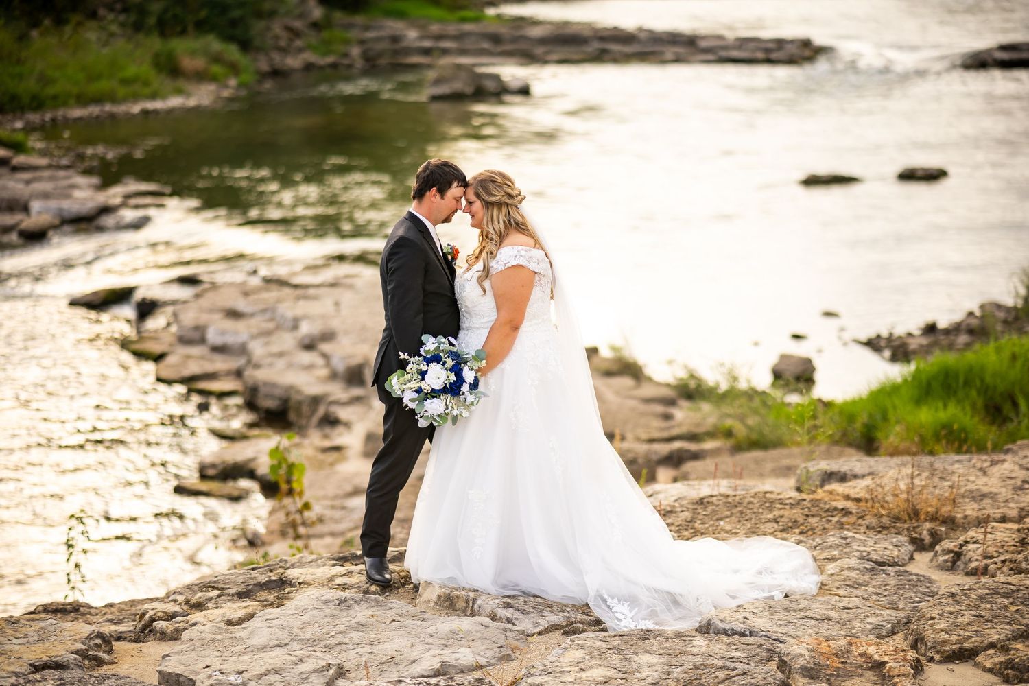Bride and groom share a romantic moment on rocky shoreline, bride holding blue and white bouquet in flowing white gown.