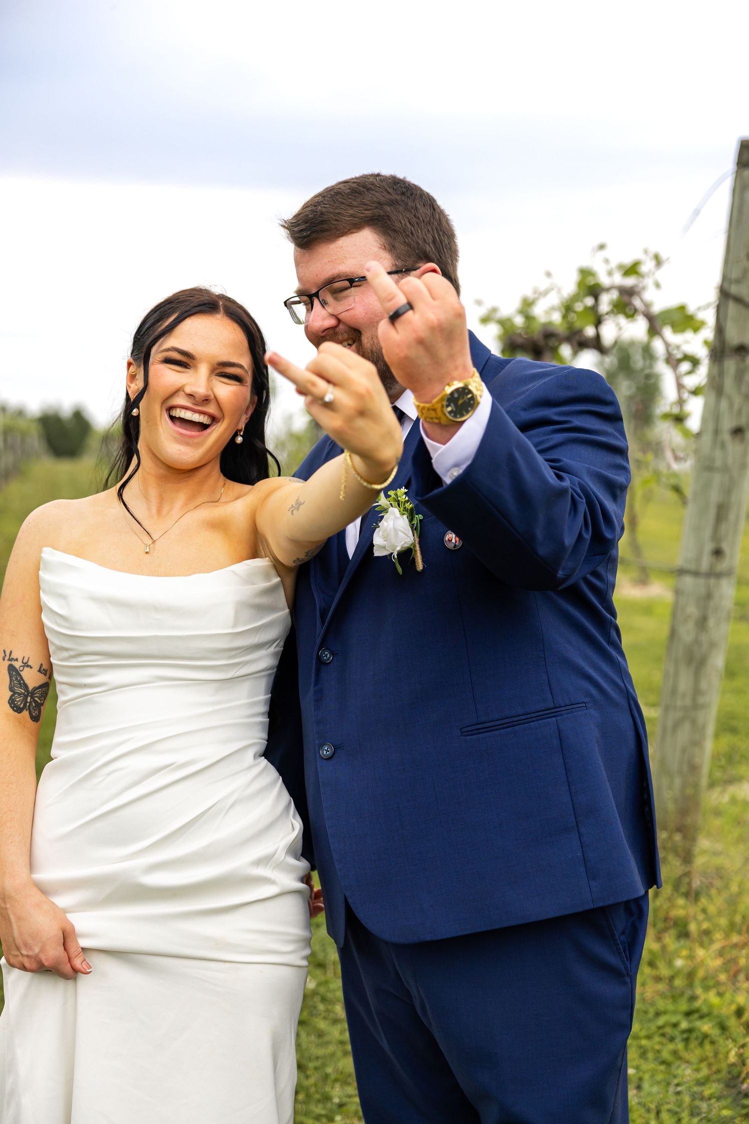 Joyful newlyweds showing off wedding rings, bride in white gown and groom in blue suit, standing in vineyard.