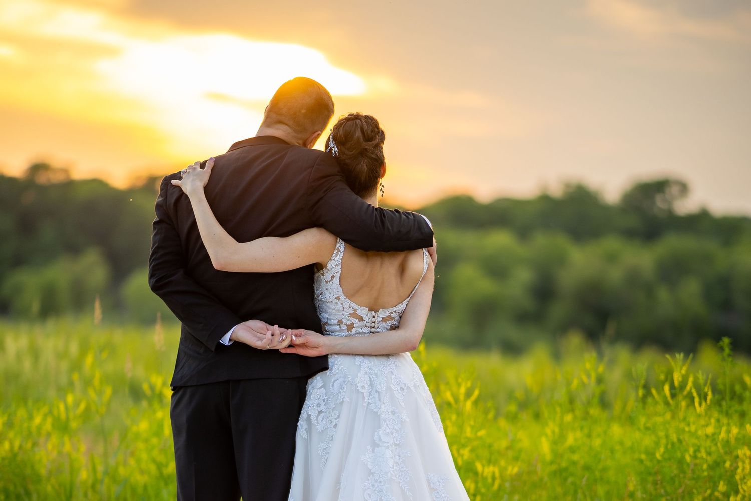 A couple embraces in a golden sunlit meadow during sunset while wearing formal wedding attire.
