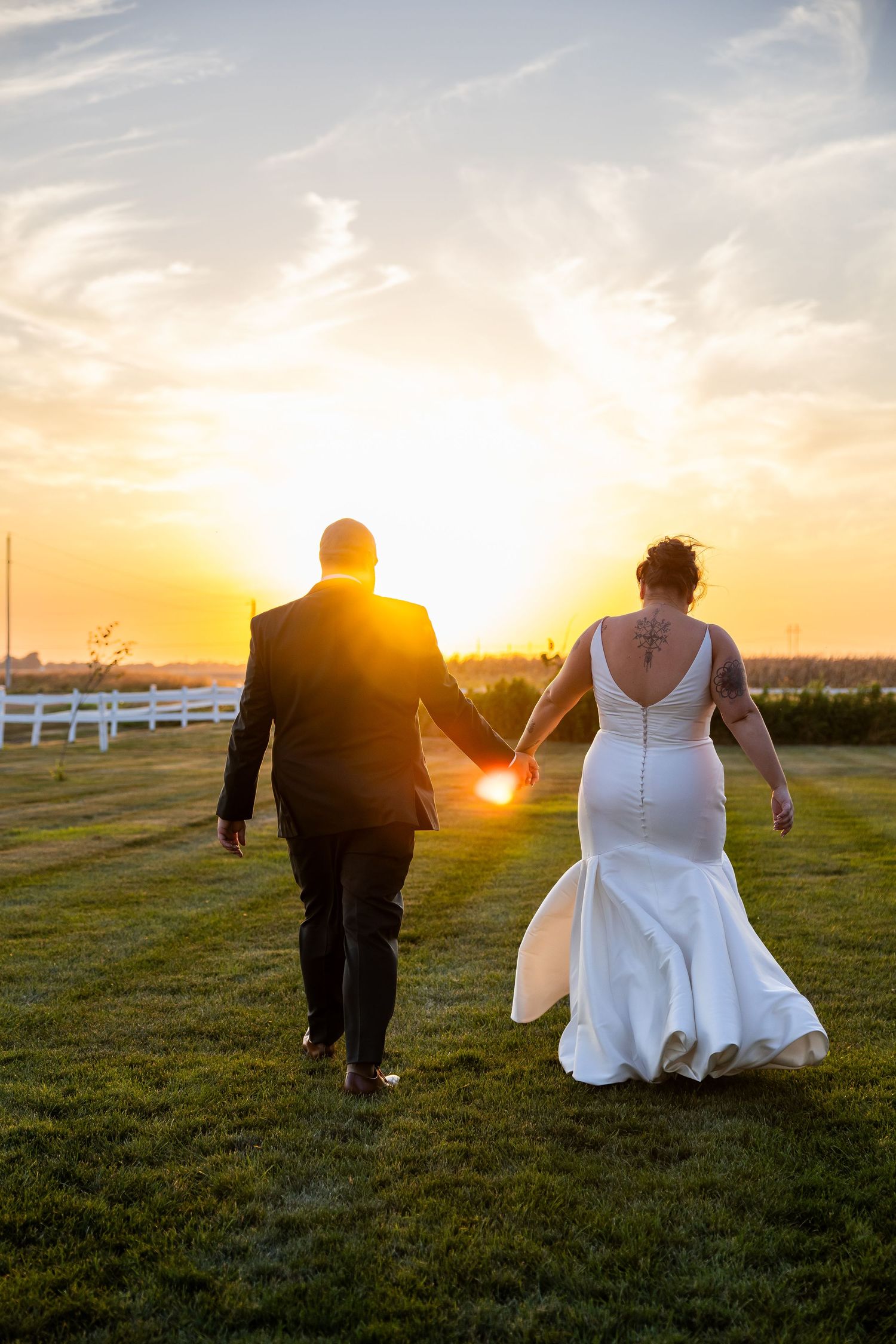 A couple walks through a grassy field at sunset with a white fence in the background, creating a romantic silhouette.