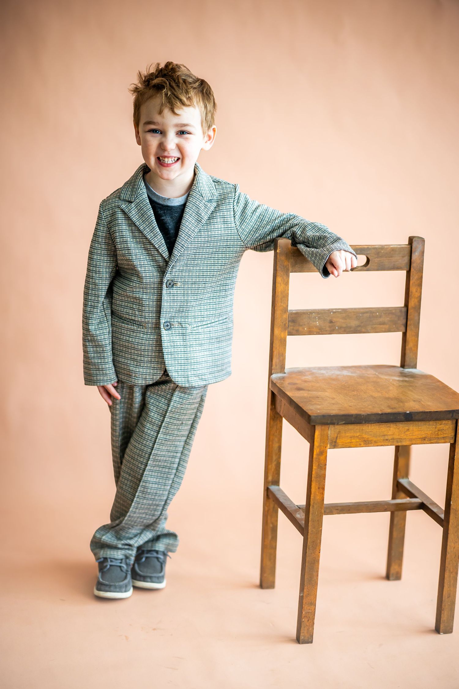 Young boy in plaid suit smiling beside a wooden chair against a peach background.