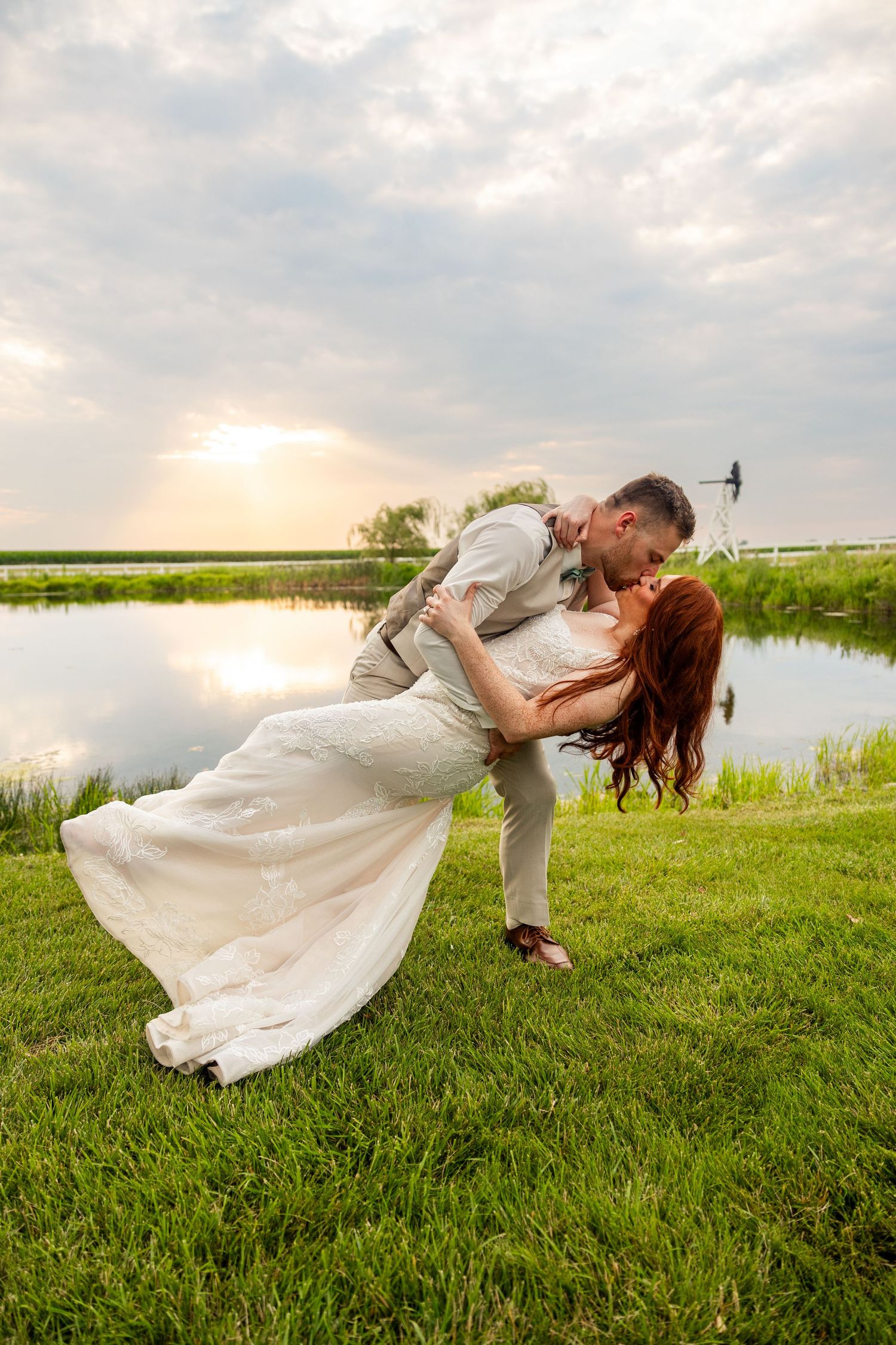Romantic couple in wedding attire share an intimate dip pose by a serene lake at sunset.