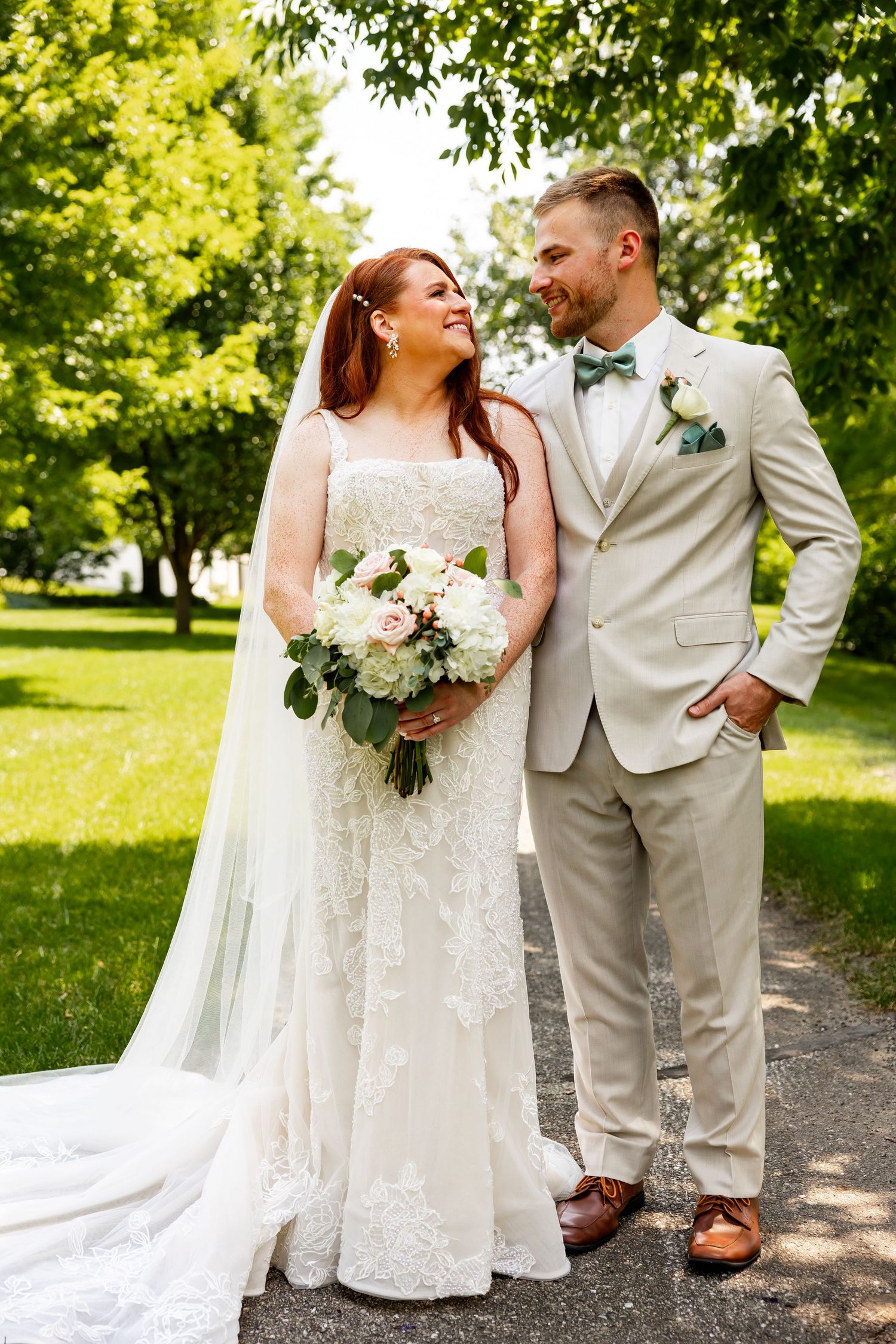 Newlywed couple laughing together outdoors, bride in lace dress and groom in light beige suit.